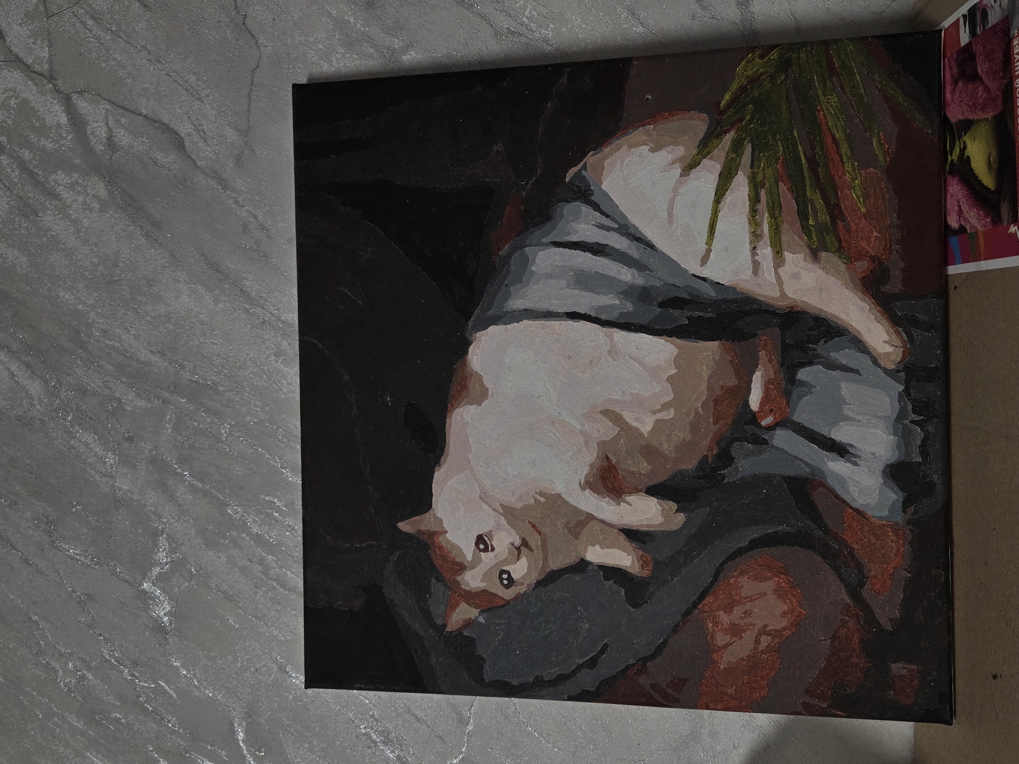 The image shows a painting of a cat lying on a surface, possibly a bed, draped with a grey fabric. The cat is mostly white with areas of reddish-brown fur. A leafy green plant is visible at the top right corner of the painting. The painting has a dark background and the overall style is reminiscent of oil or acrylic painting. The painting is displayed against a grey, marble-like wall or surface on the left side of the image. A stack of magazines or printed materials is partially visible to the right of the painting. The location is Unknown.