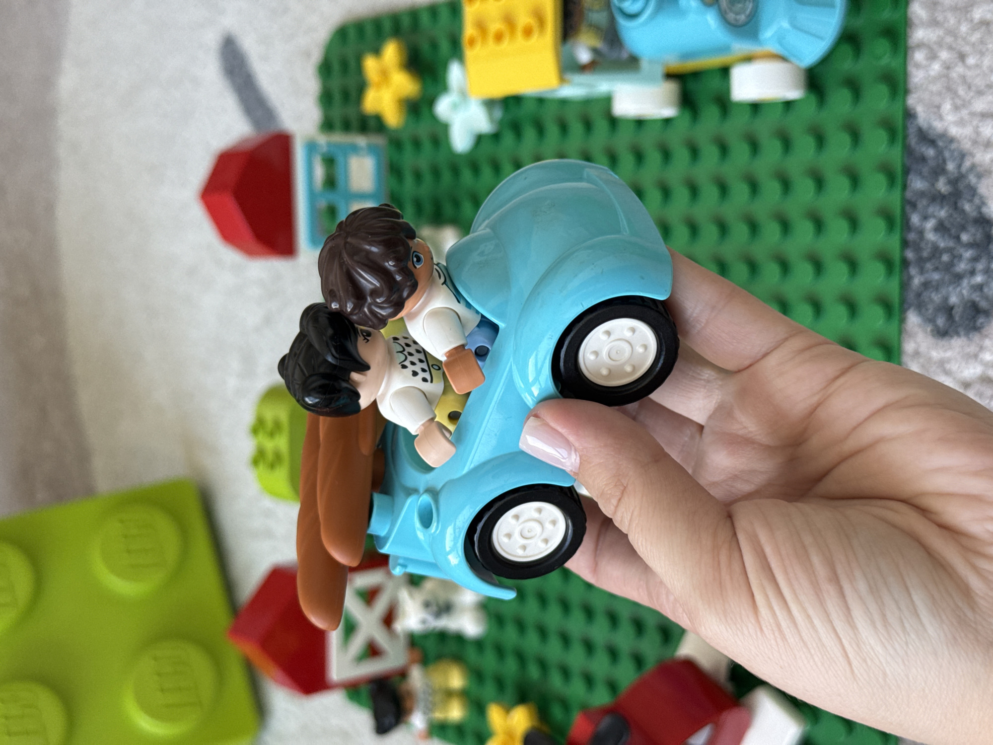A close-up, eye-level shot shows a hand holding a blue toy car with two Lego figures inside. The Lego figures are two children, one with black hair and the other with brown hair, both wearing white shirts. They are sitting in the car, which is on a green Lego base plate. In the background, there are several other Lego building blocks, including red and white houses, and yellow and white flowers. The image is well-lit, and the focus is on the car and the figures. The scene appears to be a playful arrangement of toys, likely in a domestic setting. The location is not visible in the image.