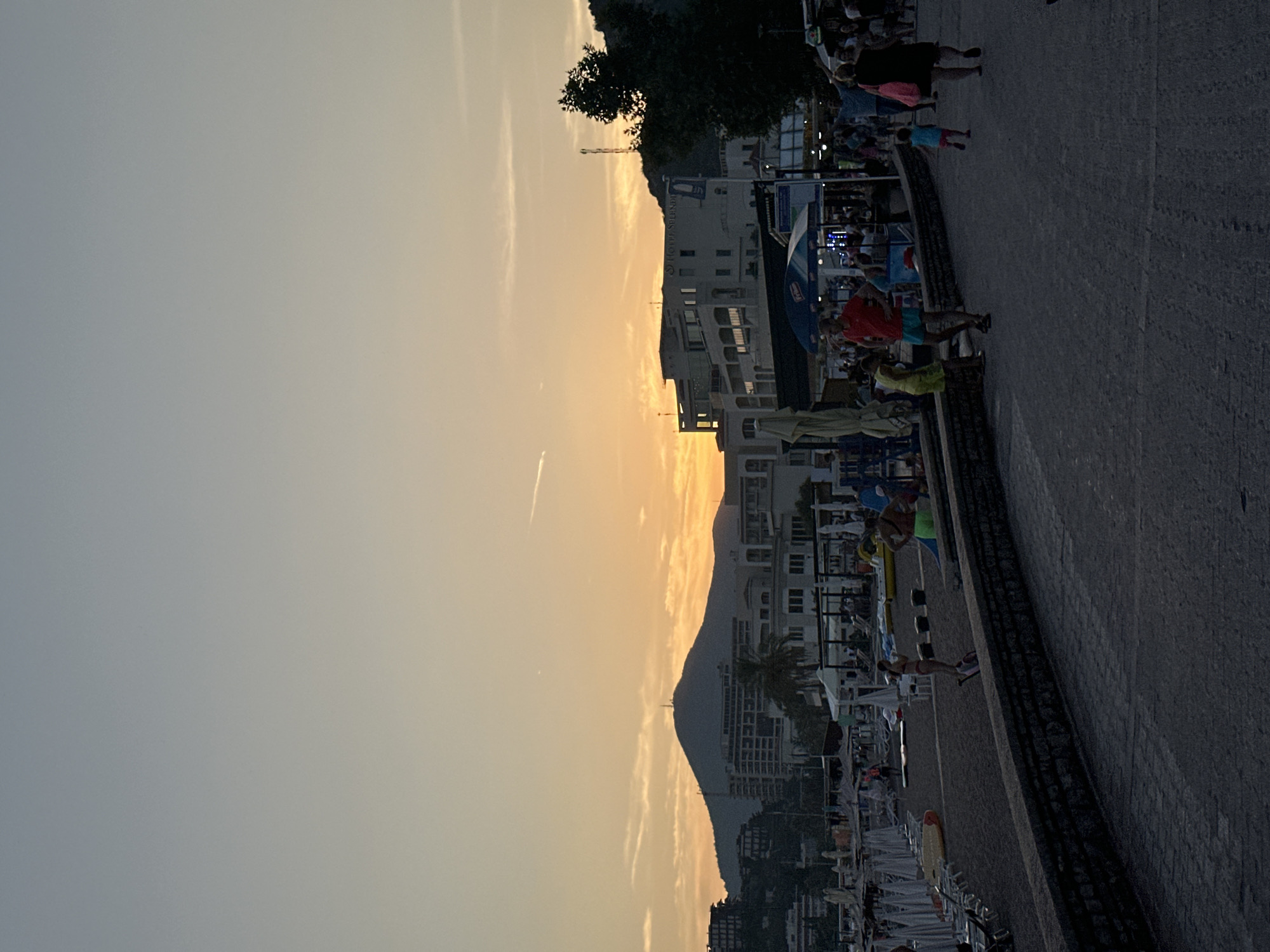 The image captures a twilight scene on a beach promenade in Boreti, Montenegro. The sky is a gradient of pale blue and soft orange, indicative of sunset. In the background, a large, dark mountain dominates the horizon, with several multi-story buildings nestled on its slopes and along the coastline. These buildings appear to be hotels and other structures typical of a resort town.
In the mid-ground, a beach is visible with numerous white lounge chairs and umbrellas, suggesting a public beach area. A stone wall separates the beach from a paved promenade where several people are walking. The people are diverse in age and attire, with some children present. One woman in a dark dress is walking away from the camera, while a young child in a blue top and pink shorts runs ahead of her. Further left, a man in a red shirt and blue shorts stands, looking down, next to a young person in a yellow garment.
The promenade itself is made of dark, textured paving stones. To the left of the promenade, some awnings and stalls are visible, indicating commercial activity. The overall atmosphere is one of a relaxed evening at a seaside location.