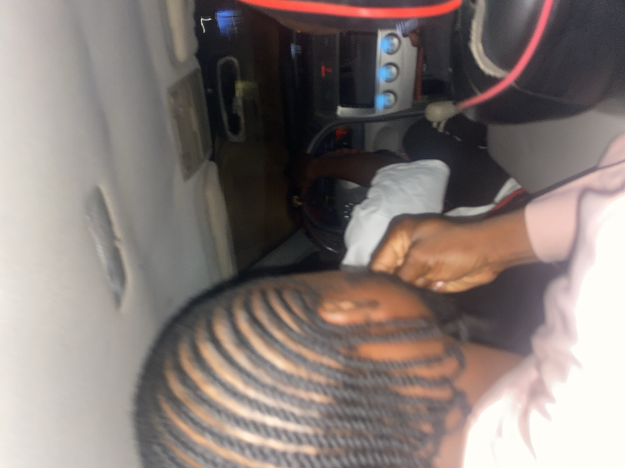 The image appears to be taken inside a vehicle. A person with braided hair is visible in the foreground. Someone's hands, possibly the driver's, are near the steering wheel. A dashboard with buttons is also visible. The location is suggested to be Jos, Nigeria, but there's no visual confirmation of the city in the image.
