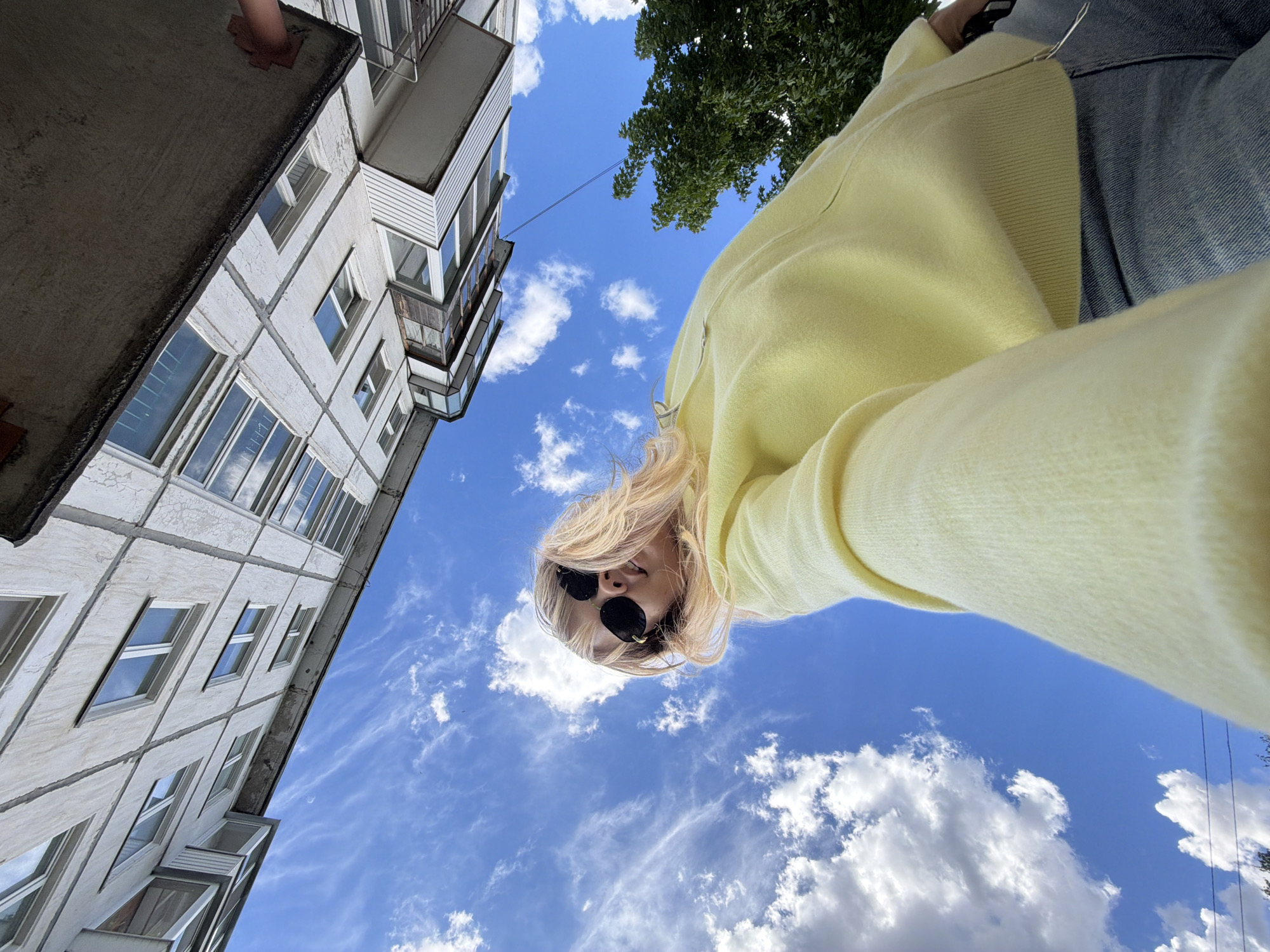 A low-angle, wide-shot photograph shows a woman with blonde hair and dark sunglasses, wearing a light yellow sweatshirt and denim jeans, standing outdoors against a backdrop of a clear blue sky with scattered white clouds. To the left of the frame is a multistory, grey-colored building with numerous windows, partially visible due to the low-angle shot. The top of a green tree is visible in the upper right corner. The photograph was taken in Chaikovsky, Russia.