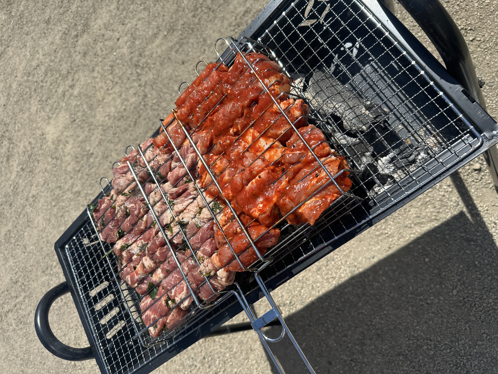 The image shows a close-up, high-angle shot of a charcoal grill loaded with meat. The grill is placed on a concrete surface, likely outdoors. Two types of meat are visible: one marinated with a red sauce and the other seasoned with green herbs. The grill has a metallic, cage-like basket containing the meat, positioned over charcoal remnants. The environment appears to be a sunny outdoor setting. There are no people directly visible in the frame. No information available to identify the location in Blagoveshchensk, Russia.