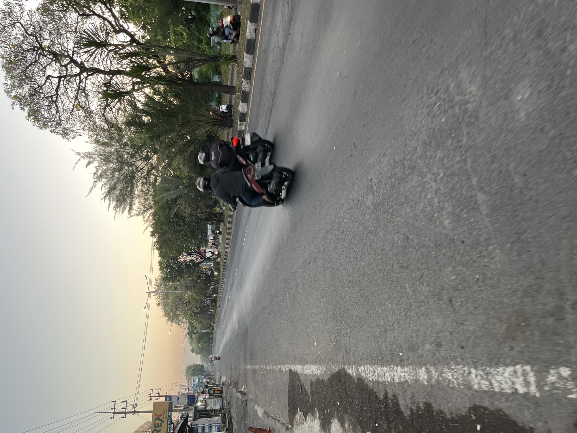 The image captures a street scene in Ngaban, Indonesia, during what appears to be dawn or dusk, given the soft, warm light illuminating the sky. The perspective is from the viewpoint of someone traveling along the road, likely on a motorcycle, evidenced by the motion blur.
In the foreground, the asphalt road dominates the lower half of the frame, marked by a faded white line on the left. To the left of the road, a sidewalk is visible with some debris and a portion of a building complex with signage, including "REX" and "ATM". A traffic cone is also present on the sidewalk.
Further down the road, several individuals on motorcycles are in motion. Two motorcycles are prominently featured in the middle ground, moving away from the viewer. The riders are wearing helmets and dark clothing, and their vehicles are slightly blurred due to movement. Beyond them, more motorcycles and a few cars can be seen in the distance, contributing to the sense of a moderately busy street.
The right side of the road is lined with trees, some with sparse foliage and others with more dense greenery, including palm-like plants. The trees and the sky, which transitions from a pale yellow to a soft orange, suggest a tropical or subtropical location. Streetlights are visible along the roadside, casting a subtle glow. The overall environment appears to be a well-trafficked urban or semi-urban area.