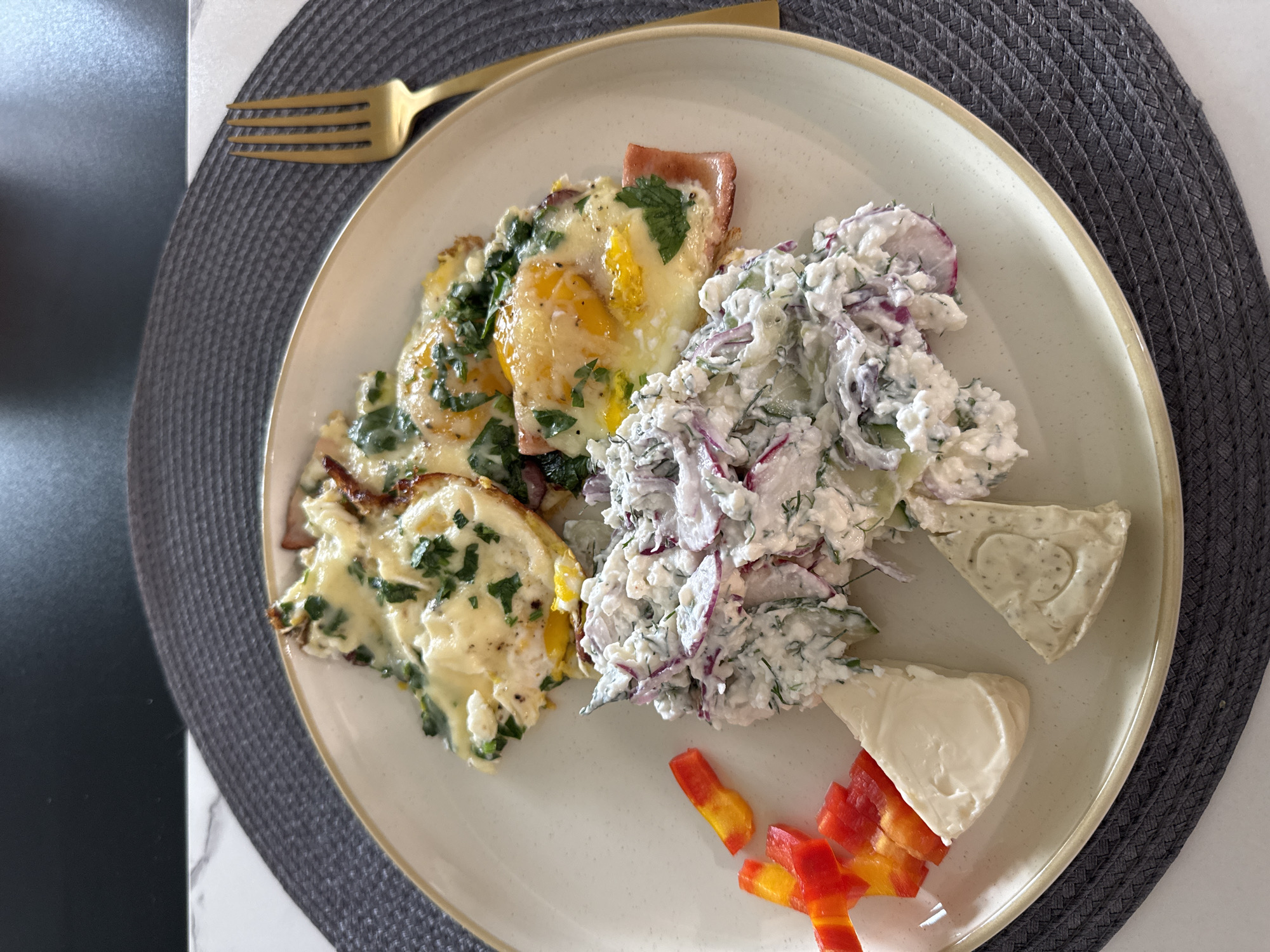 A plate of breakfast food sits on a gray placemat. The plate contains scrambled eggs with spinach and ham, a salad made with radishes and dill, pieces of red and yellow bell pepper, and two wedges of cheese. A gold fork rests to the right of the plate. The scene is set on a white marble countertop. There is no visible information to determine the location in the city of Wheeling, United States of America.