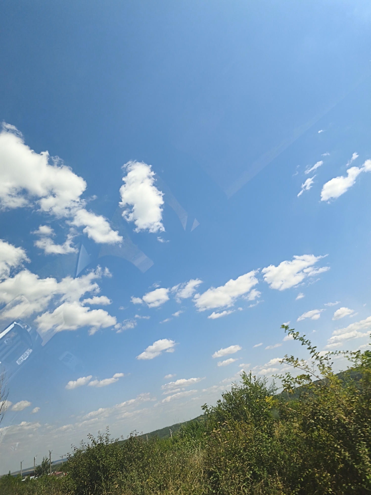 This image captures a rural landscape in Colonita, Moldova, under a clear blue sky dotted with fluffy white clouds. The foreground is dominated by lush green vegetation, including dense bushes and taller grasses. In the middle ground, a sloping hill covered in greenery is visible, with some distant structures that suggest a small settlement. A few utility poles with wires are also present, indicating the presence of infrastructure. The sky occupies a significant portion of the frame, with the clouds casting subtle shadows on the landscape below. There are reflections visible on what appears to be a window, suggesting the photo was taken from inside a vehicle. No people are visible in the scene.