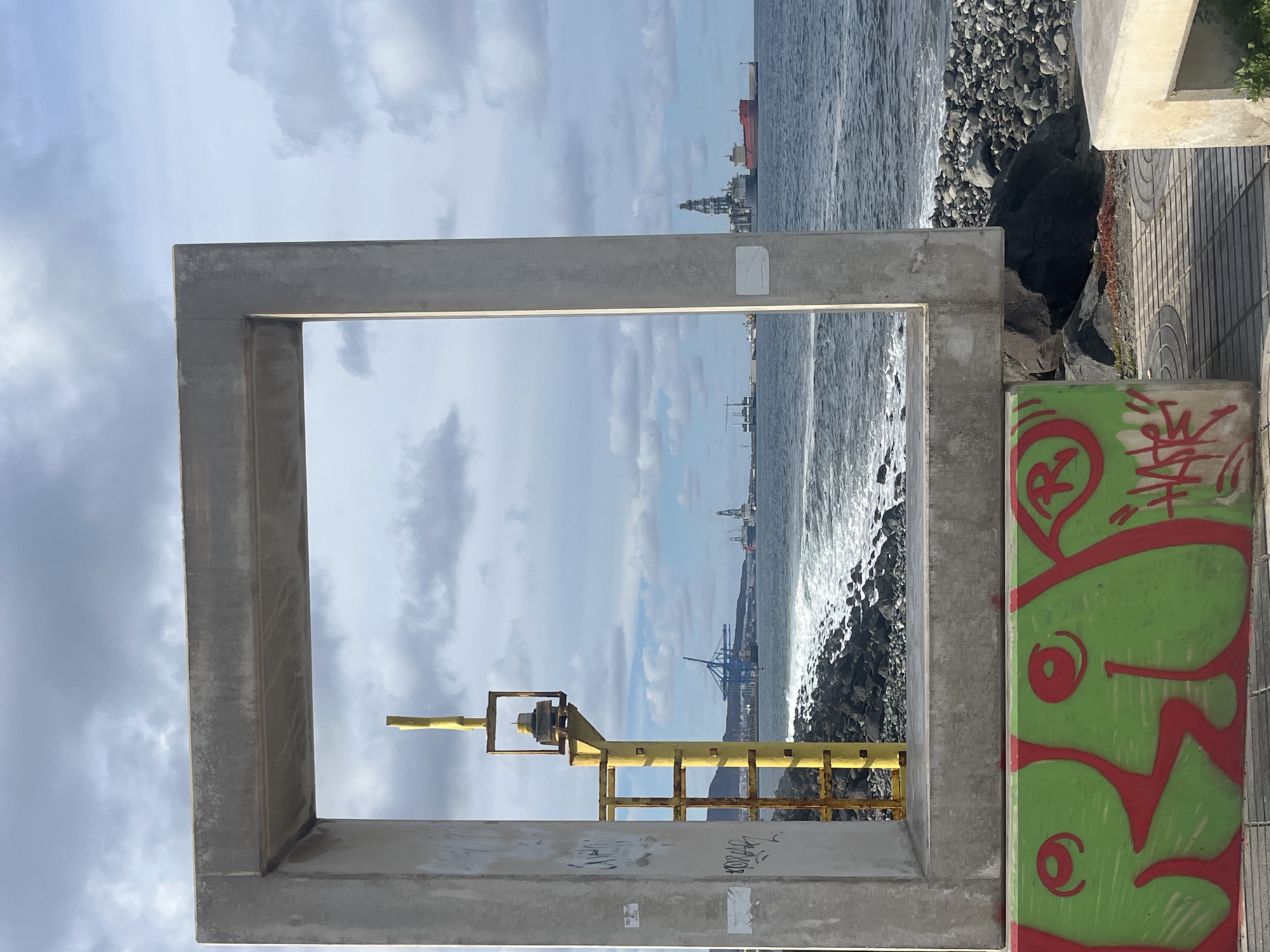 The image shows a concrete frame structure with a square opening, situated near the coast in Las Palmas, Spain. Through the frame, the view encompasses the sea, a rocky shoreline, and in the distance, maritime infrastructure including cranes and a ship. A yellow, possibly metal, structure with a ladder-like appearance stands within the frame's view, closer to the viewer. To the right of the concrete frame is a concrete wall with graffiti art in green and red. The sky is overcast with clouds.