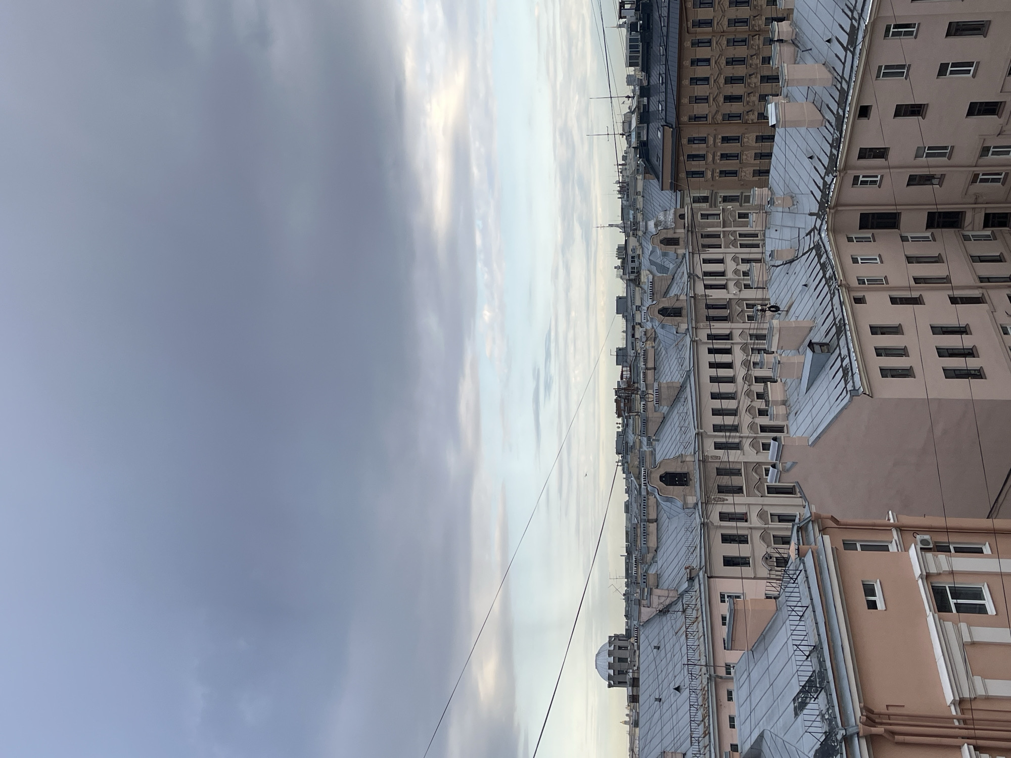 The image displays a wide, high-angle view of a dense urban landscape in Saint Petersburg, Russia, under an overcast sky. The scene is dominated by a vast expanse of building rooftops extending towards the horizon, with a cloudy sky occupying the upper half.
The sky features a mix of grey clouds, varying from darker, thicker formations on the left to lighter, brighter sections towards the right, indicating an overcast or partly cloudy daytime.
Below the skyline, a multitude of multi-story buildings are tightly packed together. The architecture suggests a historical European city, with a variety of facades and roof styles. Many buildings in the foreground and midground have pitched roofs, often covered in grey or metallic materials, adorned with numerous chimneys, vents, and occasional small dormer windows. The closest buildings exhibit light pink or peach-colored facades with rows of rectangular windows, some featuring decorative trims. Further in the distance, buildings appear in various shades of beige, light brown, and grey. Several thin, dark utility wires or cables are visible stretching horizontally across the middle distance against the sky. No specific activities or individuals are discernible in the static cityscape. The overall lighting suggests it is daytime, without strong shadows to indicate a specific time. No visible text is present.