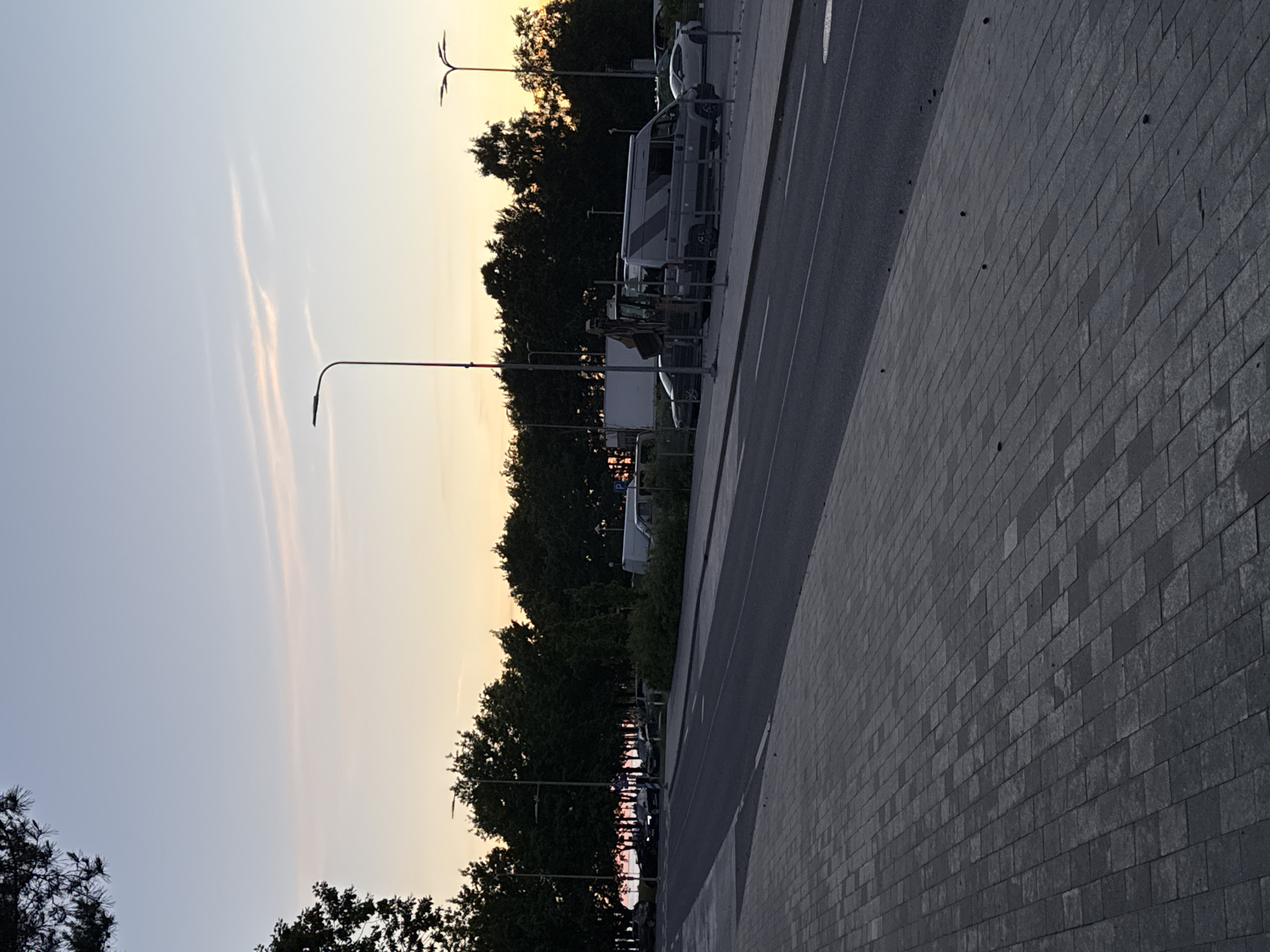 The image depicts a street scene in Tallinn, Estonia during sunset. The sky is a pale blue with wisps of pink and orange clouds. Trees line the background, and a paved sidewalk is in the foreground. Several vehicles are parked along the street, including a grey van, a white van, a construction excavator, and several cars. Streetlights are visible throughout the scene. The overall atmosphere suggests a quiet evening in a residential or commercial area.