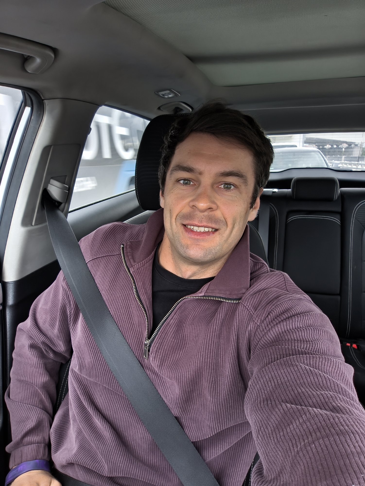 The image is a selfie of a man wearing a purple corduroy jacket and a black shirt, with a seatbelt fastened across his chest. He is smiling at the camera. He is inside a car. The car has black leather seats with white stitching. Through the windows, a blurred cityscape is visible. Based on the blurry signage and the architectural style of the visible buildings, the location might be Moscow, Russia. There are other vehicles visible outside the car window.
