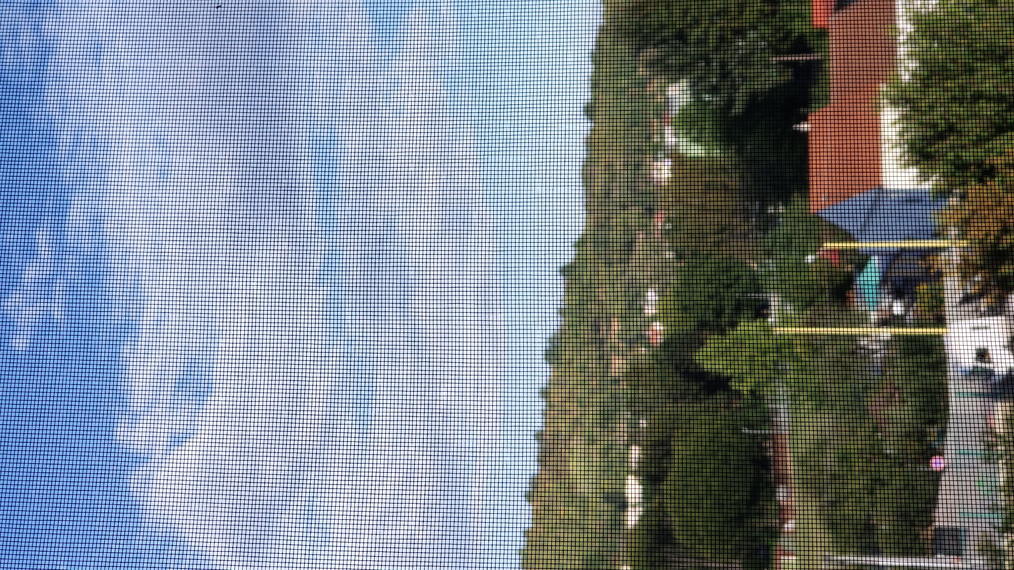 This image depicts a suburban landscape viewed through a window screen, with a clear blue sky dotted with white clouds overhead. Below the sky, rolling green hills are covered in dense foliage, interspersed with glimpses of houses with reddish-brown roofs. The foreground reveals more vegetation, including trees and some houses. Notably, a white structure, possibly a vehicle, is visible in the lower portion of the frame, alongside what appears to be goalposts, suggesting a sports field or recreational area. The presence of these elements points towards a residential area with some amenities. Without specific landmarks or identifying features, it is not possible to pinpoint the exact location within Prešov, Slovakia.