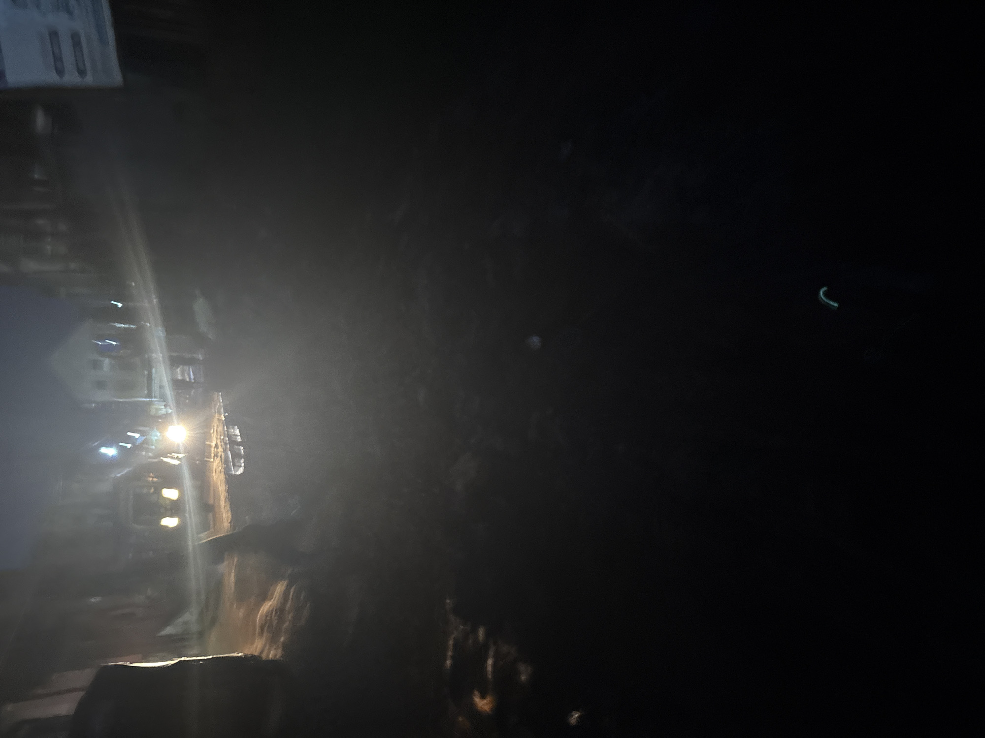 The image depicts a dimly lit street scene at night, possibly in Onitsha, Nigeria, characterized by low visibility and a hazy atmosphere. In the background, several vehicles with illuminated headlights are visible, suggesting movement or stationary presence on the road. The lighting from the vehicles creates a somewhat blurred and diffused effect, typical of a night shot with limited light sources. Structures that appear to be buildings line the street, but their details are obscured by the darkness and atmospheric conditions. The ground surface seems to be wet or reflective, possibly indicating recent rain or the presence of puddles, which reflect the available light. A faint, blue-green glow is visible in the lower right portion of the image, its source and nature unclear. The overall impression is one of a nocturnal urban environment with challenging lighting conditions.