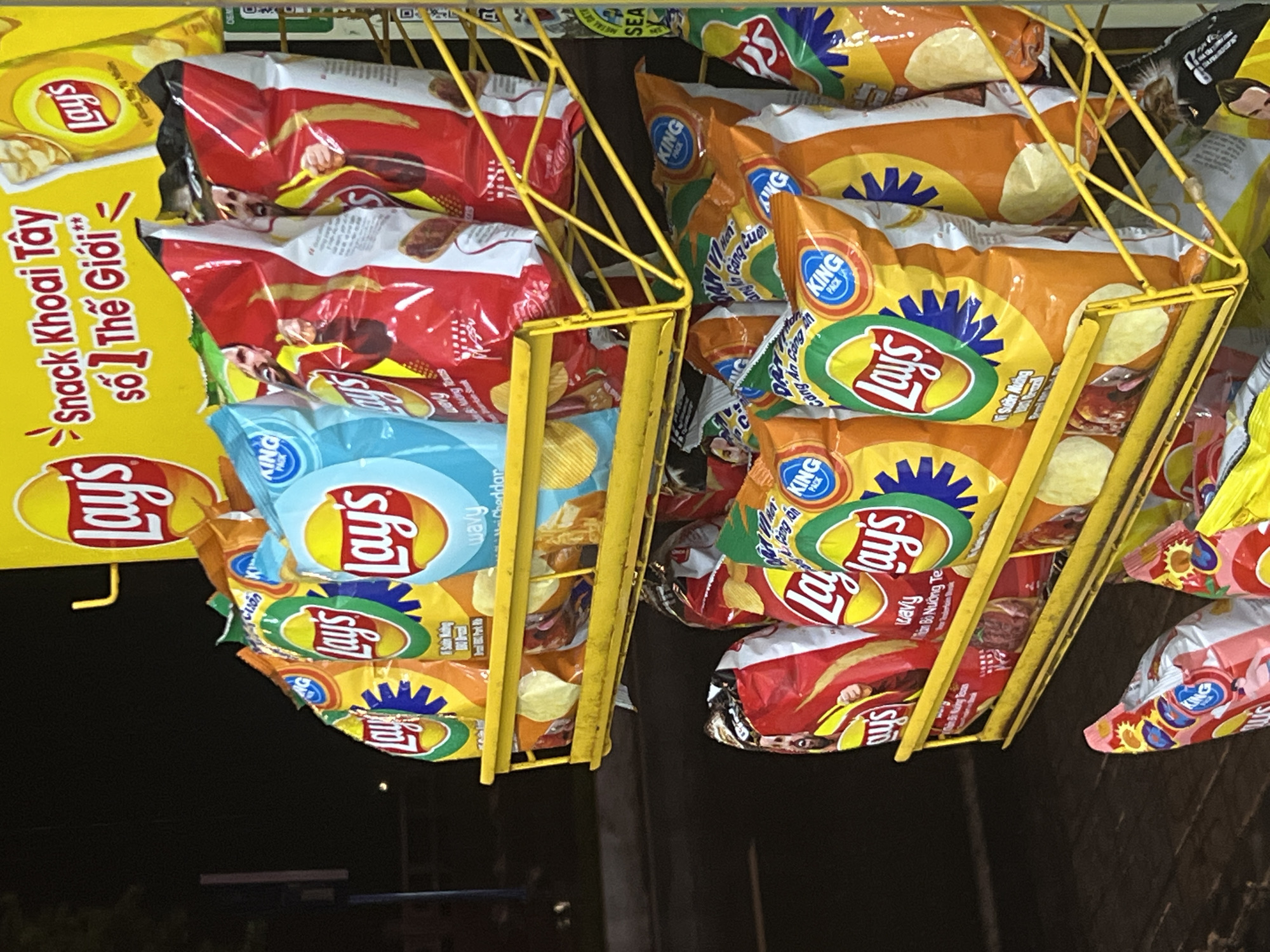 Captured from a low angle, the image depicts two yellow metal racks filled with Lay's brand potato chip bags, suspended against a dark backdrop. The rack on the left features bags of Lay's chips, including flavors labeled "Snack Khoai Tây" and "Cheddar." The right rack contains Lay's and King Pack chip bags with flavors written in Vietnamese such as "Sườn Mương." A sign marked with "Snack Khoai Tây số 1 Thế Giới" is partially visible on the left. The scene appears to be a retail display in Bac Nha Trang Ward, Vietnam.