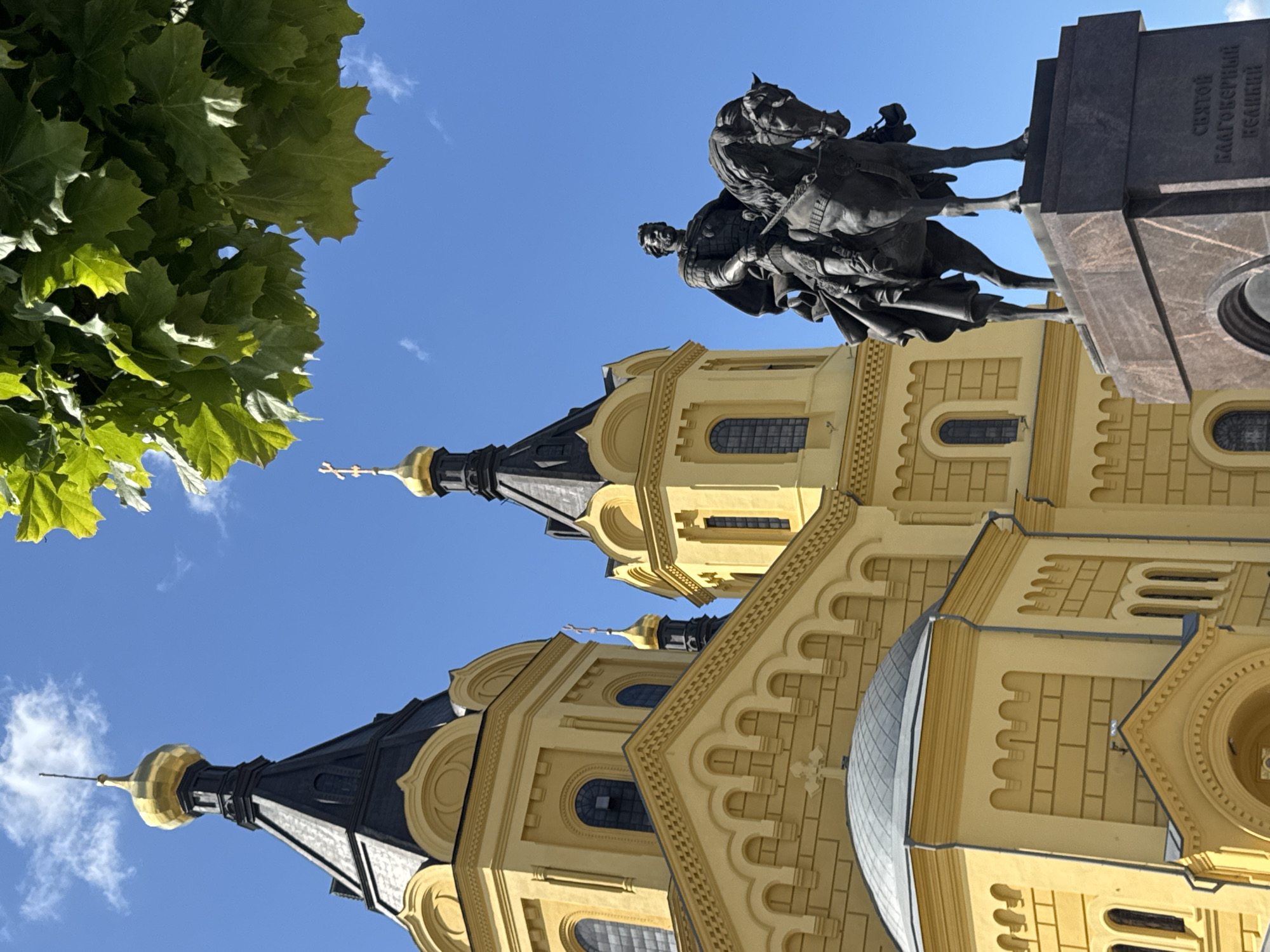 The image captures a close-up view of the Cathedral of Alexander Nevsky in Nizhny Novgorod, Russia, with an equestrian statue in the foreground. The yellow facade of the cathedral, adorned with architectural details and topped by a black roof with a golden dome, is visible against a clear blue sky. Sunlight illuminates the building, casting shadows. To the right of the cathedral, a dark bronze equestrian statue depicts a man in armor on horseback, mounted on a large, dark granite pedestal. The statue is positioned such that the cathedral serves as a backdrop. Lush green leaves from a tree frame the top right portion of the image, providing a natural element to the scene. The base of the statue has some Russian text inscribed on it.