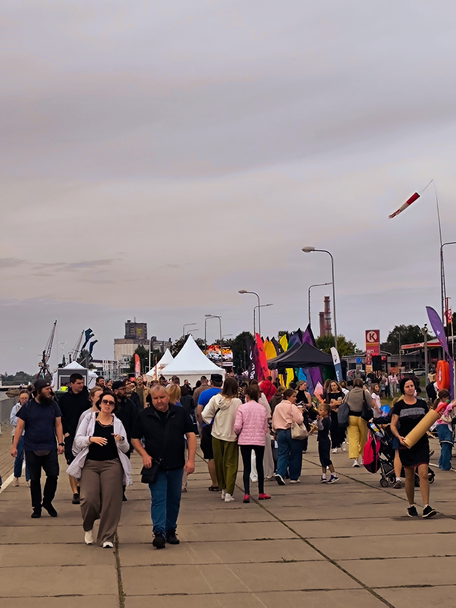 The image depicts a bustling outdoor event in Riga, Latvia, possibly a festival or market, along a waterfront promenade. The foreground features a crowd of people walking on a paved walkway. Several individuals are walking towards the viewer, some in small groups.
In the midground, there are white tents set up, suggesting vendors or event activities. Colorful flags of various sizes and designs are prominently displayed, some attached to poles, others appearing to be part of displays. A black tent with rainbow-colored fabric is visible. In the background, industrial elements are present, including cranes and a tall building with a distinctive silhouette, possibly a silo or factory, indicative of Riga's port or industrial areas. A red and white windsock is visible in the upper right corner, indicating wind. A Circle K gas station sign is also visible in the distance, suggesting an urban setting near a transportation route. The sky is overcast with muted tones. The overall impression is a lively public gathering taking place on a day with diffused lighting.