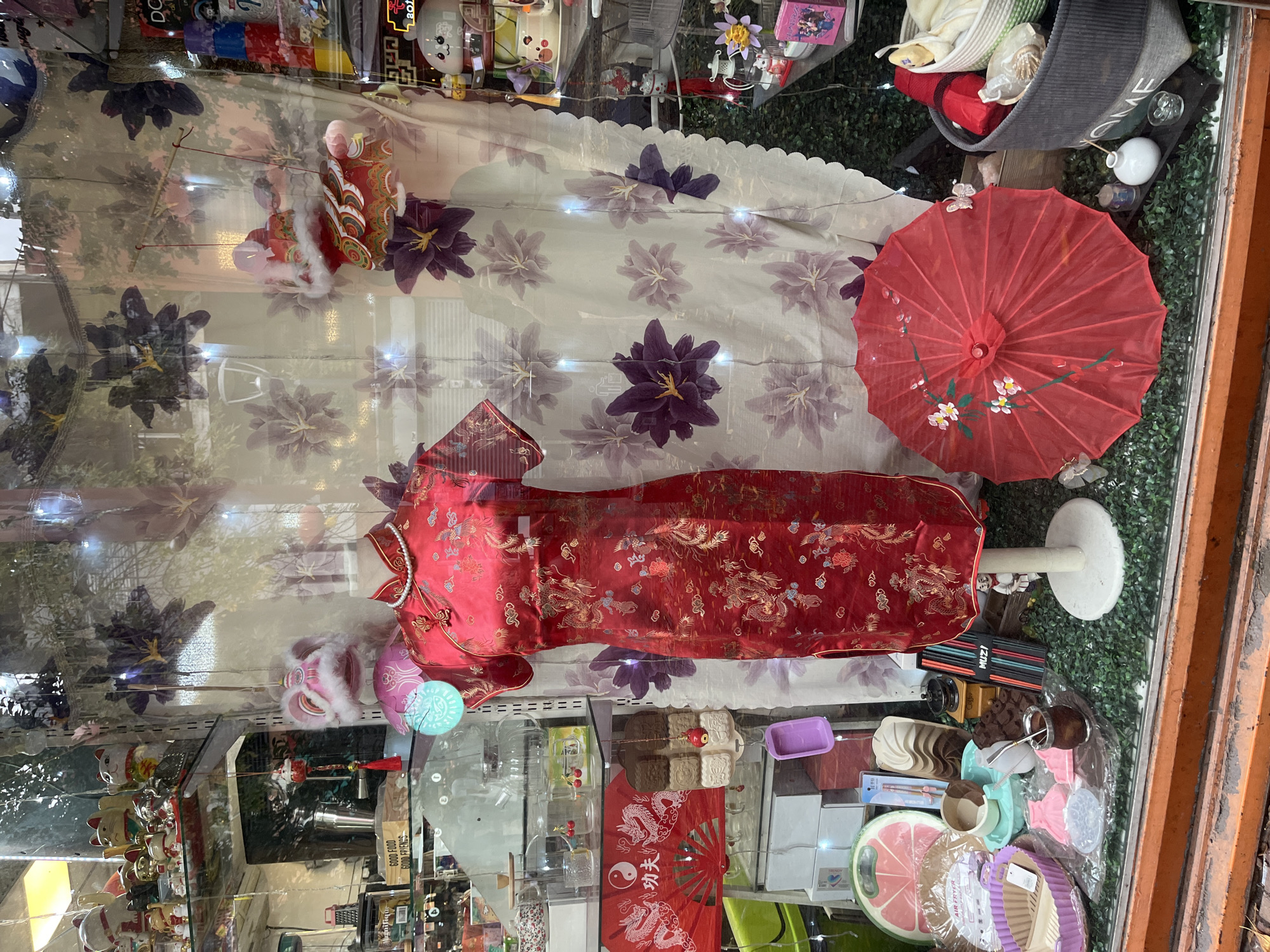 This image captures a vibrant display window of a store in Buenos Aires, Argentina, seen through a glass pane with noticeable reflections of the outside environment, including greenery. The display is densely packed with a variety of objects, many of which are associated with Asian culture and celebration, particularly Chinese.
The central and most prominent item is a richly embroidered red Qipao (cheongsam) dress, adorned with intricate gold dragon and floral patterns, displayed on a white mannequin torso with a pearl necklace. To its left, a decorative red and gold lion dance head with white fur is visible, and further down, a smaller pink lion dance figure adds to the festive theme. On the right side of the display, a bright red parasol (umbrella) featuring delicate white and pink cherry blossom paintings is open.
Behind these main elements, a white fabric backdrop with large purple and lighter-toned floral patterns is illuminated by small, scattered string lights, creating a soft glow.
The lower section of the display, closer to the viewer, is filled with a diverse collection of items, suggesting a blend of cultural artifacts and general household goods. These include traditional Chinese mooncake molds in brown and white, a red folding fan with a gold dragon design and the visible Chinese characters "功夫" (Gōngfu - Kung Fu) alongside a Yin-Yang symbol. Interestingly, a traditional South American mate gourd with a bombilla is also present, reflecting the local context. Other items include various silicone molds (pink, purple, green), a large grapefruit-slice-shaped cutting board or platter, a manual coffee grinder, and a black basket with the word "HOME" printed on it. Several lucky cat (Maneki-neko) figurines in different sizes and colors are also displayed, along with small glass containers holding cherries and other packaged goods, one prominently labeled "AIR FRYER." Faint text like "GOOD FOODS GOOD FRIENDS" is visible on a shelf edge.
The overall scene is a static retail display, likely intended to attract customers with its festive and varied offerings. The vibrant colors and cultural items evoke a sense of tradition and celebration.