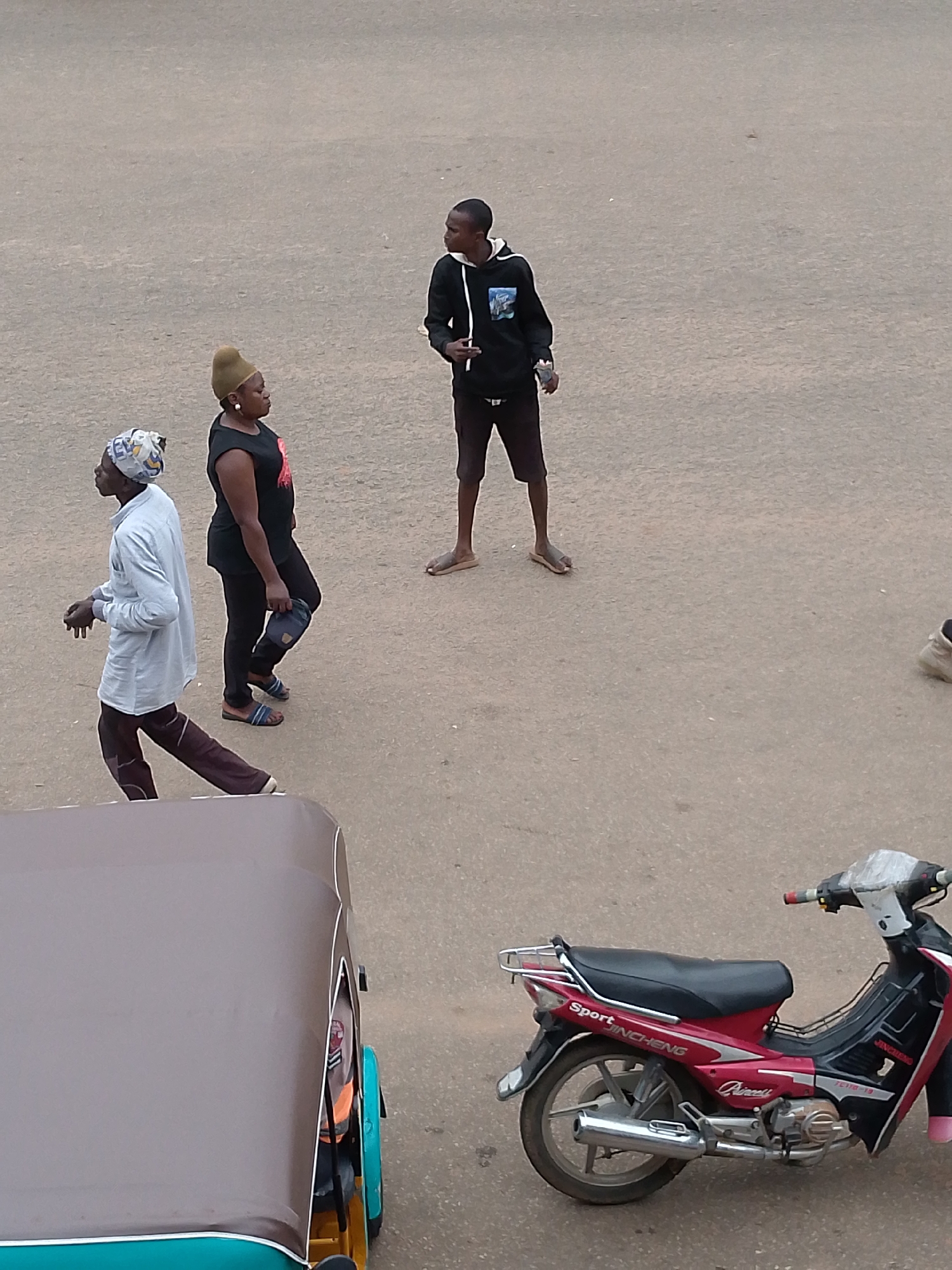 In Bukuru, Nigeria, on a paved road, three individuals are walking. On the left, a person wearing a light blue long-sleeved shirt and brown trousers walks with their hands clasped. To their right, a woman with dark clothing and a beige headwrap walks, carrying a blue cap in her hand. Further to the right, a young man in a black hoodie and dark shorts stands facing towards the left. In the foreground on the right, a red and black motorcycle is parked. In the bottom left corner, a portion of a brown and teal three-wheeled vehicle is visible. The ground is a muted, grayish-brown asphalt.