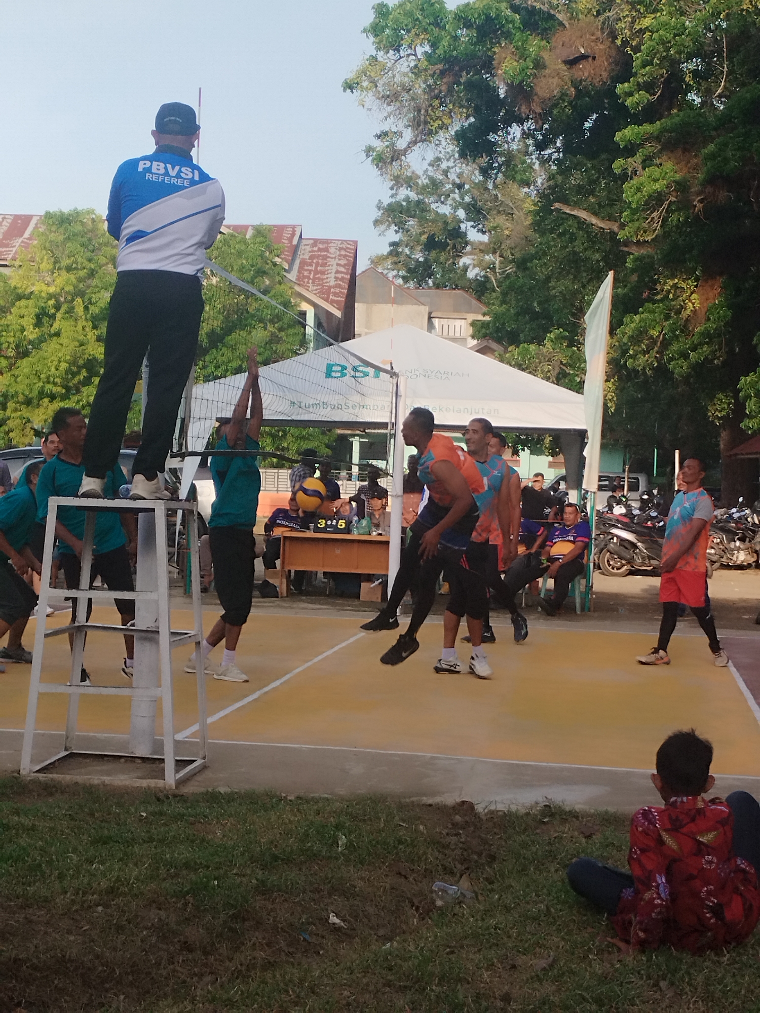 The image depicts an outdoor volleyball match in Lhokseumawe, Indonesia. The scene captures several individuals engaged in the sport, with a referee standing on an elevated platform. The court is marked with yellow and white lines, and a volleyball net is visible. In the background, there are trees, buildings, and parked motorcycles. One person, wearing a red shirt with a floral pattern, is sitting on the grass in the foreground, facing away from the camera. The time of day appears to be late afternoon, given the lighting and the long shadows cast on the court. The overall atmosphere suggests a community sporting event.