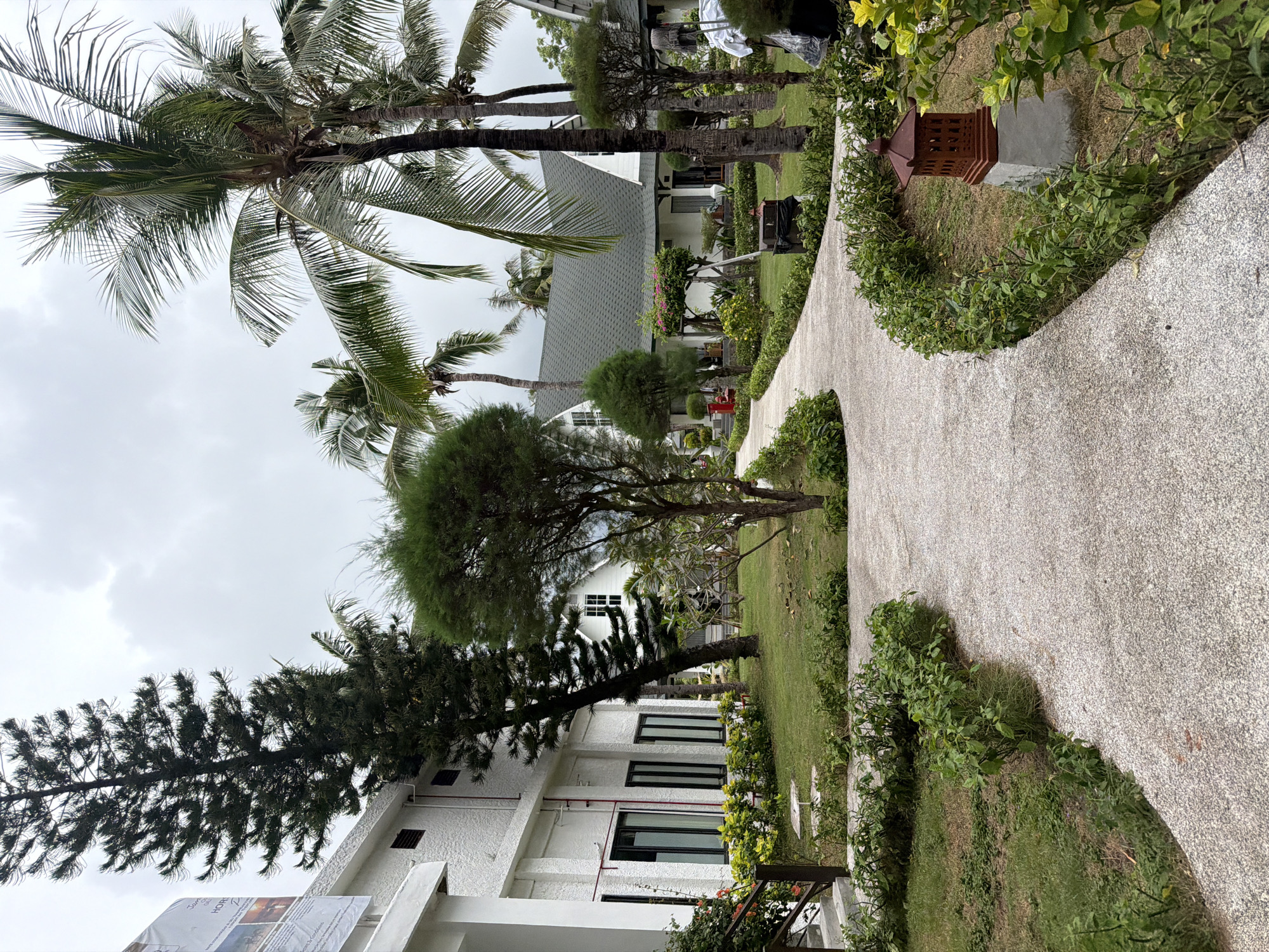A serene, overcast day unfolds at a tropical resort in Unknown, Indonesia. A winding gravel pathway meanders through lush greenery, separating two white buildings with dark-framed windows. One building, closer to the viewer on the left, displays a large banner with text. Beyond the pathway, palm trees and other tropical foliage create a vibrant backdrop, framing a quaint white guesthouse with a dark roof. A small, ornate brown lantern sits beside the path, adding a touch of rustic charm to the tranquil scene. The overall atmosphere is one of peaceful retreat, characteristic of a tropical vacation destination.
