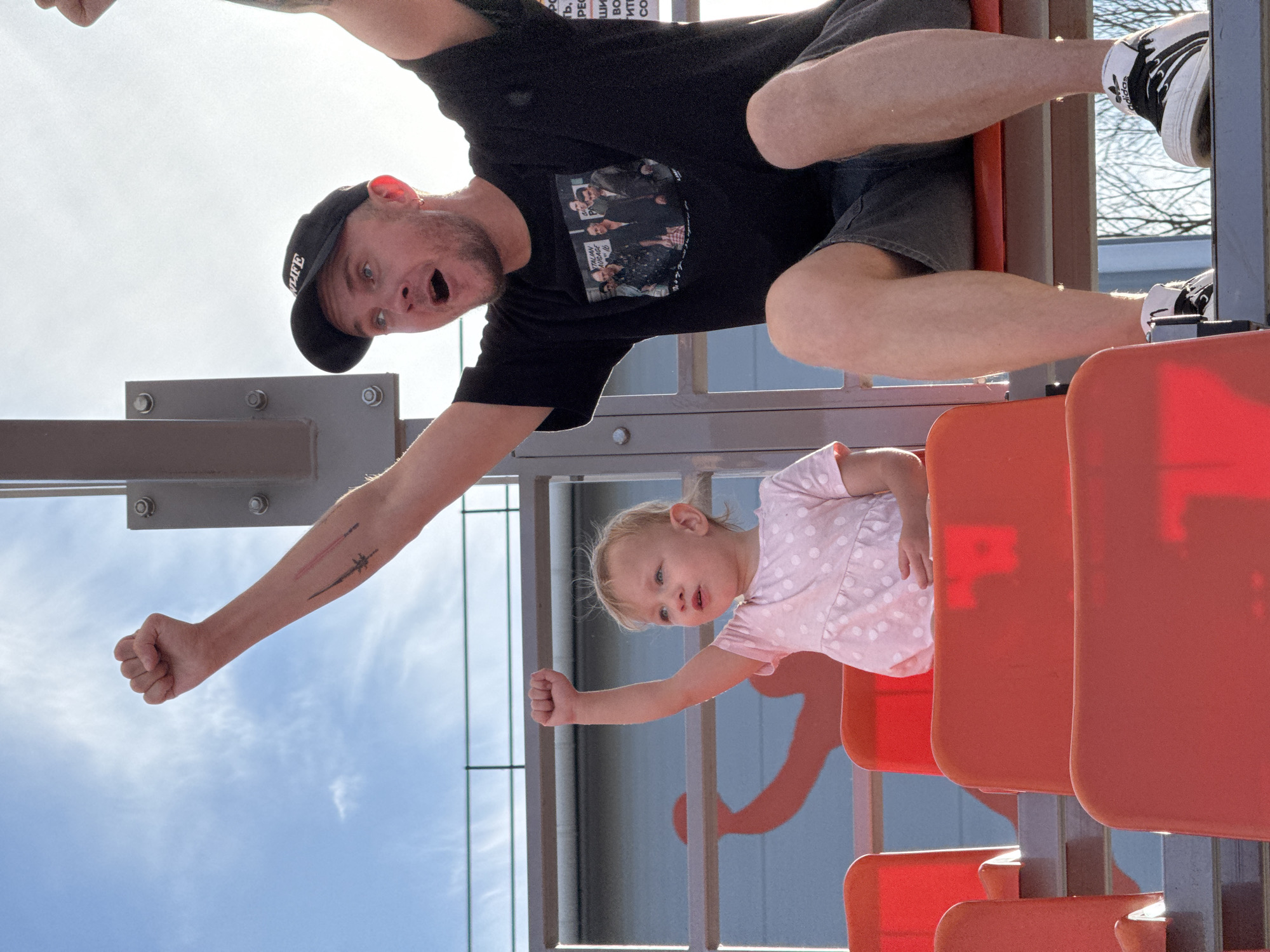 Two people, an adult male and a young child, are sitting in orange stadium seats under a bright, partly cloudy sky. The adult male, on the right, is wearing a black t-shirt with a picture on it, grey shorts, a black baseball cap, and white Adidas sneakers. His arms are raised in the air. The child, on the left, is wearing a pink polka dot dress and has one arm raised. They appear to be at an outdoor sporting event or concert. The image seems to have been taken in Novocherkassk, Russia, as indicated by the text on the building behind them, which includes Russian Cyrillic letters.