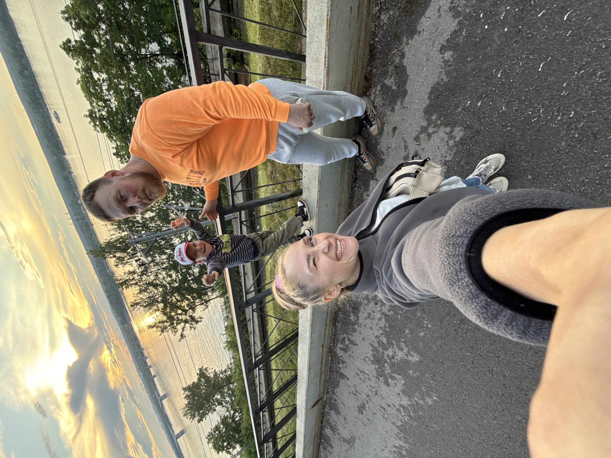 The image shows a family of three, a man, a woman, and a young child, taking a selfie with a scenic background of a river, bridge, and trees. The setting appears to be a promenade or elevated walkway, possibly near the Kama River in Perm, Russia. The woman is in the foreground, smiling and winking at the camera. The man is standing behind her, leaning on a railing, and looking towards the camera. The child is positioned between them, with arms outstretched and a playful expression. The sky is illuminated with the warm glow of a sunset, casting a golden light on the water. The overall mood is one of joy and togetherness.