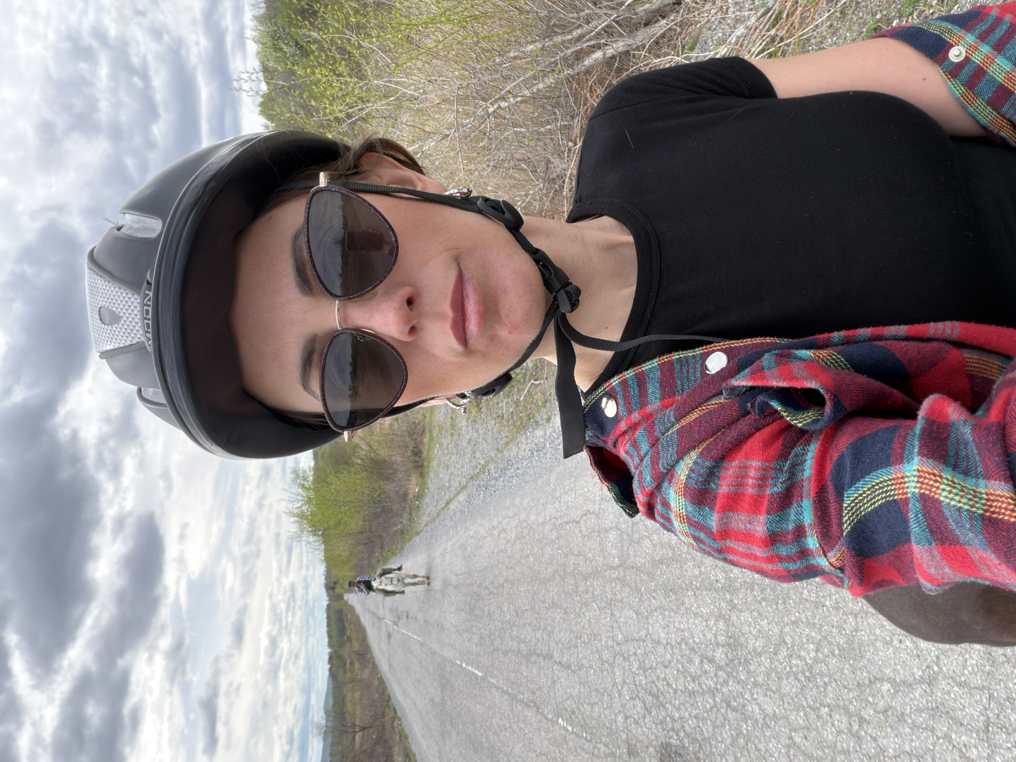 A selfie taken on a paved road with a person on a bicycle in the distance. The person taking the photo is wearing a black T-shirt, a plaid shirt, sunglasses, and a bicycle helmet. The road is cracked and appears to be in a rural area. The sky is overcast. No information about the location is explicitly available in the image.