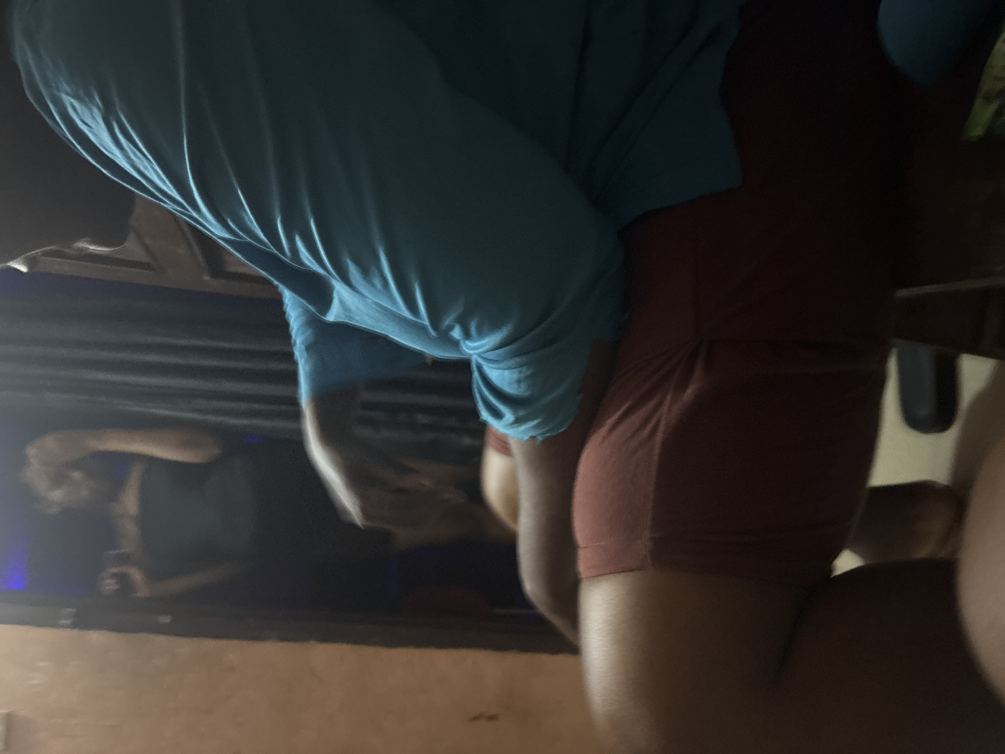 The image depicts a scene within a room in Onitsha, Nigeria. In the foreground, a person is seated, wearing a blue long-sleeved shirt and red shorts. Their body is angled away from the camera, with their left arm resting on their lap and their right hand visible. The background shows a dimly lit doorway with a person standing in it, holding a dark object, possibly a phone, and appearing to be looking at it. The person in the doorway is wearing a black strapless top. The room's ambiance is dark, with ambient light filtering from the doorway and a light-colored wall visible on the left side of the frame. Curtains are present in the background. The overall impression is of a casual, indoor setting.