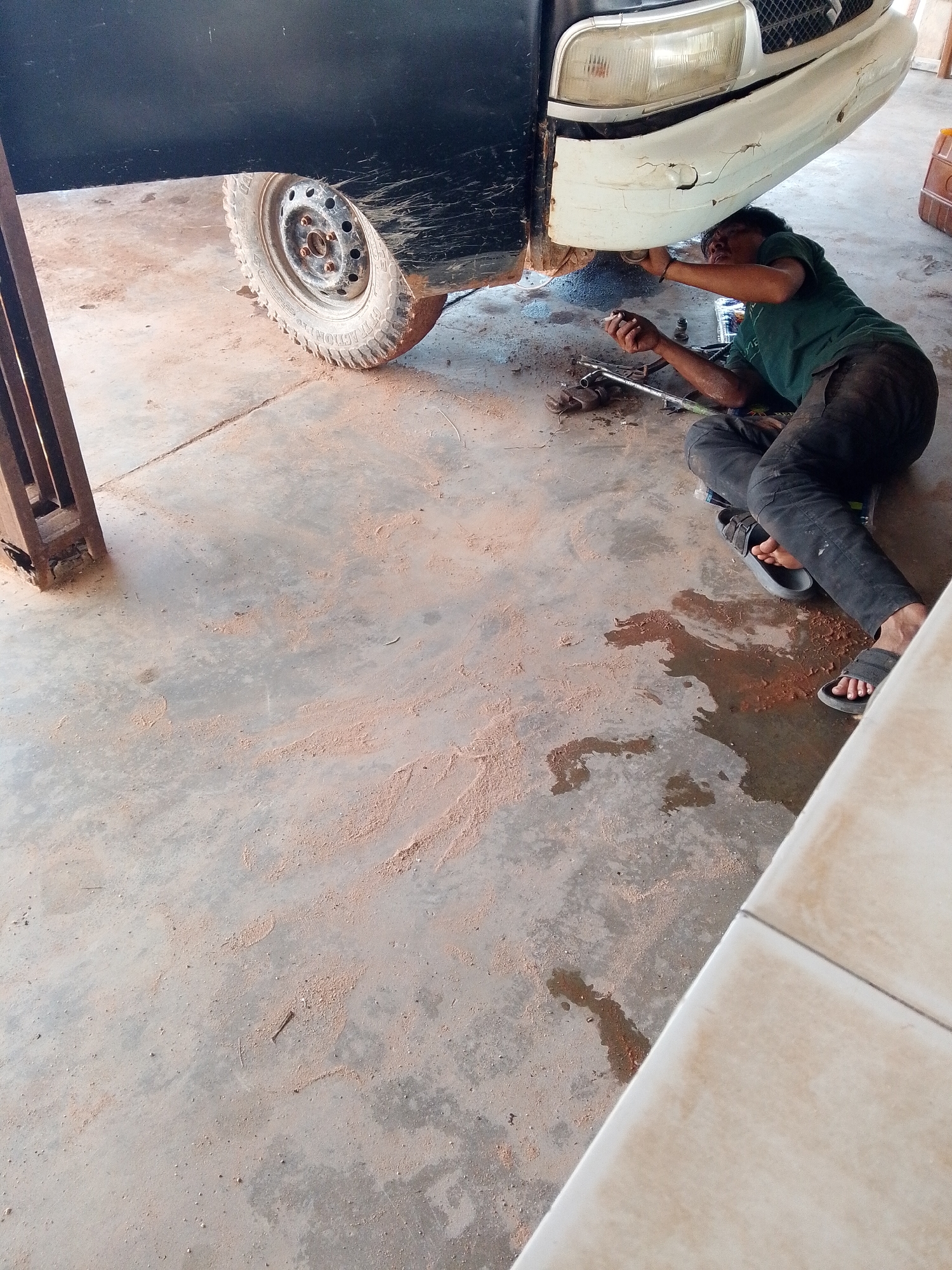 A person wearing a green t-shirt and dark pants is lying on their back under the front of a white vehicle, working on the underside of the car with tools. The vehicle is partially positioned inside what appears to be a garage or workshop, with a black section of the vehicle's body visible above the front wheel. The floor is concrete, marked with what looks like dirt, dust, and some liquid spills. The area is a garage located in the city of Bukit Ketok, Indonesia. A car lift is visible in the left part of the scene.