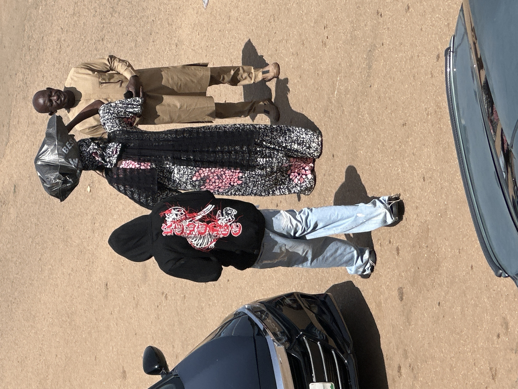 In Bukuru, Nigeria, a scene unfolds on a dusty street. Three individuals are present. To the left, a person wearing a black hooded sweatshirt with a white and red skeleton graphic on the back and light blue jeans stands facing away from the camera. To the right, a man in a tan, long-sleeved tunic and trousers, with dark skin and a bald head, looks towards the center. Between them, a person draped in a patterned black and white fabric, with a pink floral motif visible, carries a large, black plastic bag on their head. The background is a flat, sandy surface, indicative of an outdoor, possibly unpaved, area. Shadows are cast on the ground, suggesting a bright, sunny day. A black car is partially visible on the left side of the frame. Another car's hood is in the foreground at the bottom of the image.