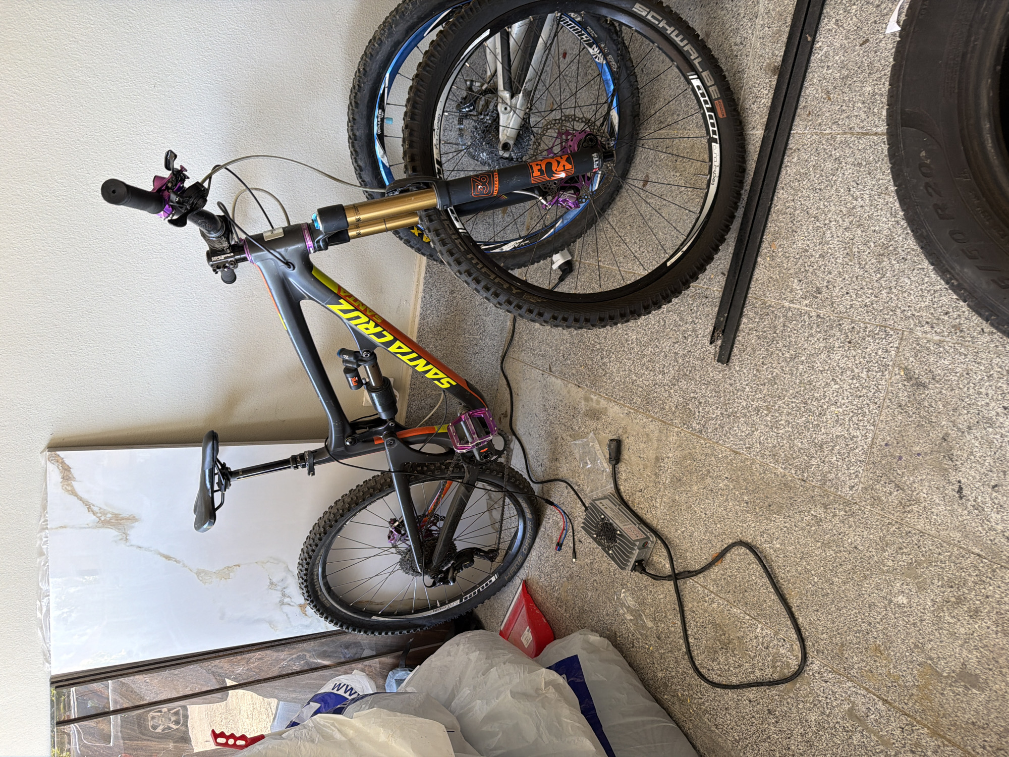 The image shows a cluttered indoor space, possibly a garage or storage area. A Santa Cruz mountain bike with purple accents is the central subject, leaning against a wall. A spare tire is stacked behind the bike. To the right of the bike are a charger and various items covered with white plastic bags. Additionally, a stack of tiles are visible against the left wall. The location in Asenovgrad, Bulgaria, cannot be determined from the provided image.