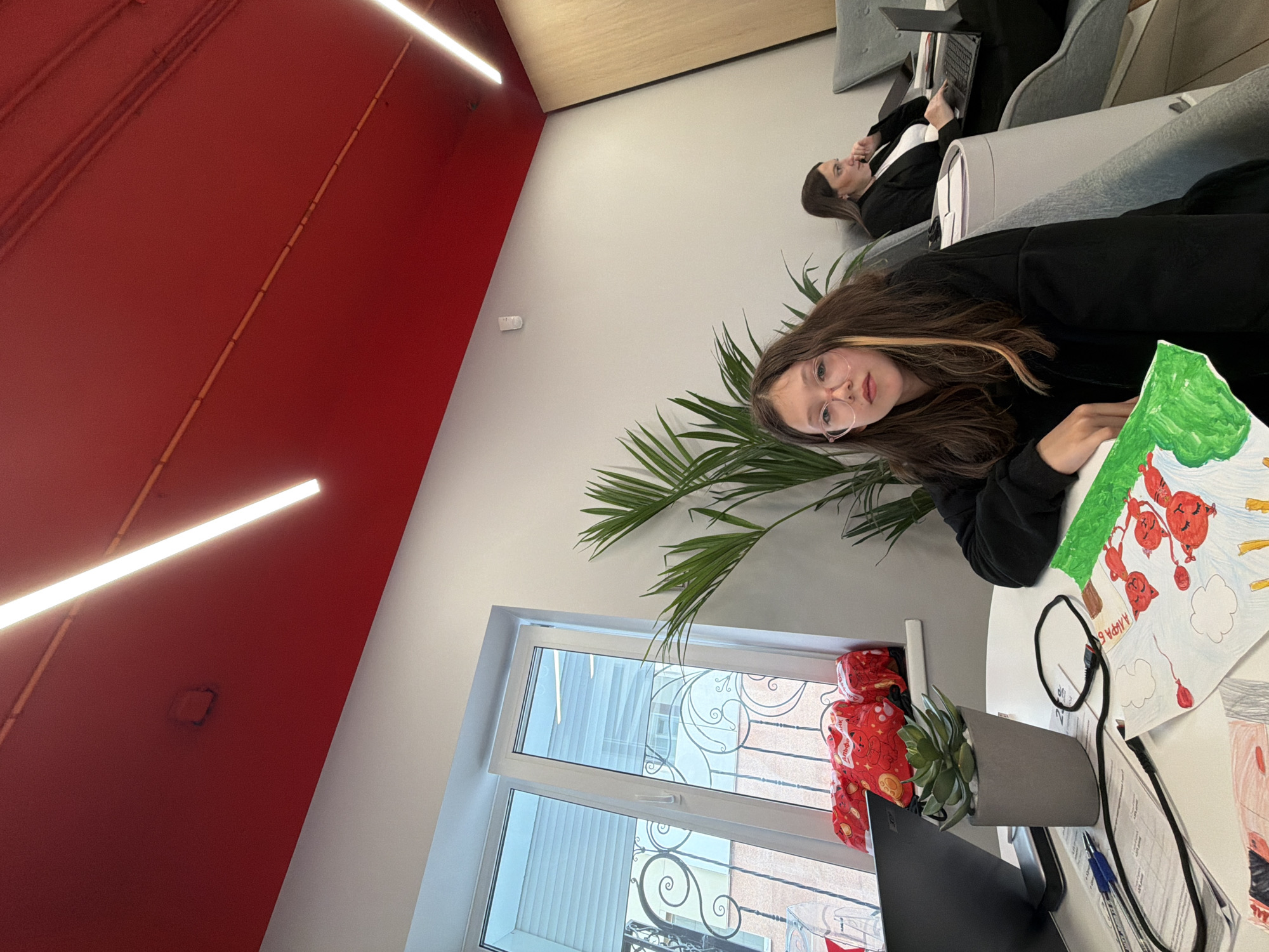 The scene is indoors, likely an office space. A girl wearing glasses and a black jacket sits at a white table, looking at the camera. She has a drawing of what appears to be balloons. Next to her is a potted plant and a laptop. In the background, another woman is seated in a grey chair, using a laptop. There's a large indoor plant behind the girl. A window is visible, revealing a building facade. The walls are white and red, with overhead fluorescent lighting. There is no definitive evidence in the image to pinpoint the exact location as Birobidzhan, Russia.