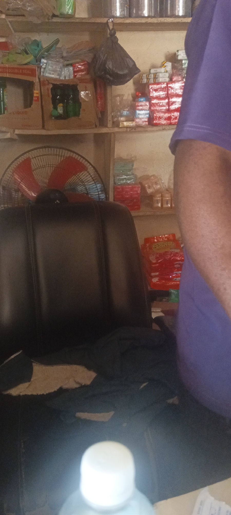 The image appears to be taken inside a small store or shop in Bokkos, Nigeria. There is a black office chair in the foreground, partly covered by a dark-colored jacket with a light-colored lining. Behind the chair, a red and black fan is visible. In the background, shelves are stocked with various goods, including stacks of red and white packaged items, bottles in cardboard boxes, and other miscellaneous items. A person wearing a purple shirt is partially visible on the right side of the frame. The environment suggests a small retail business setting. A bottle is partially visible at the very bottom.