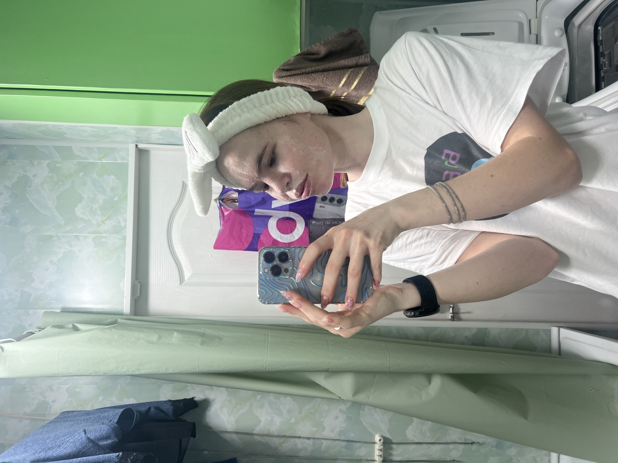 A young woman is taking a selfie in a bathroom. She is wearing a white t-shirt, a white headband with bunny ears, and a black smartwatch. She is holding a blue cell phone with a textured case. Her face appears to have a skincare product applied. The bathroom has green walls, a white door, and a light green shower curtain. A washing machine is partially visible in the background. There are shopping bags hanging on the door. The location may be Chaikovsky, Russia.