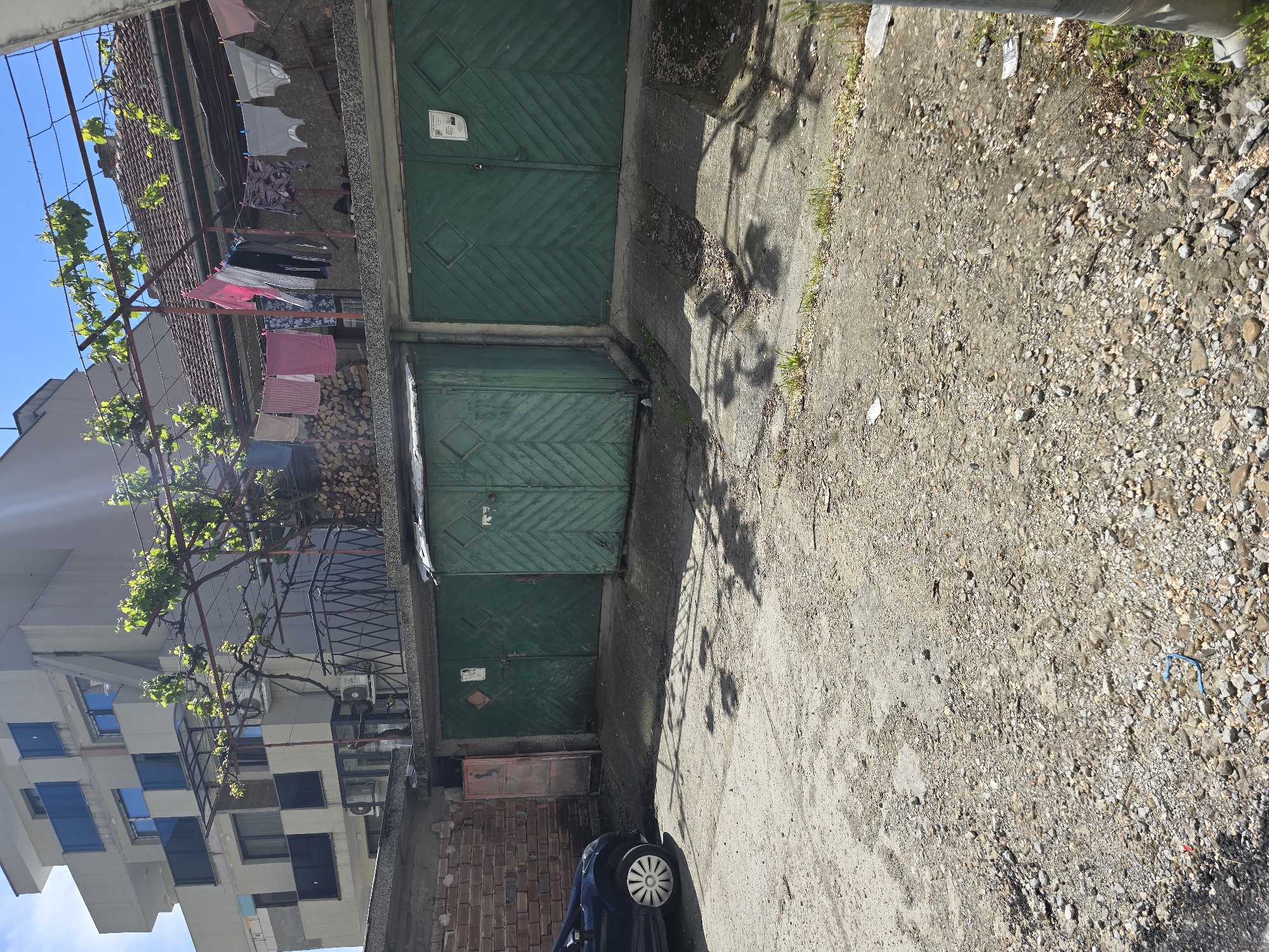 This image captures a scene in an urban location, possibly in Unknown, Unknown. The foreground is dominated by a mixed surface of concrete and gravel. There is an old garage with three green wooden doors. Laundry is hanging above them. A small car is parked at the end of the shot. Buildings are visible in the background.