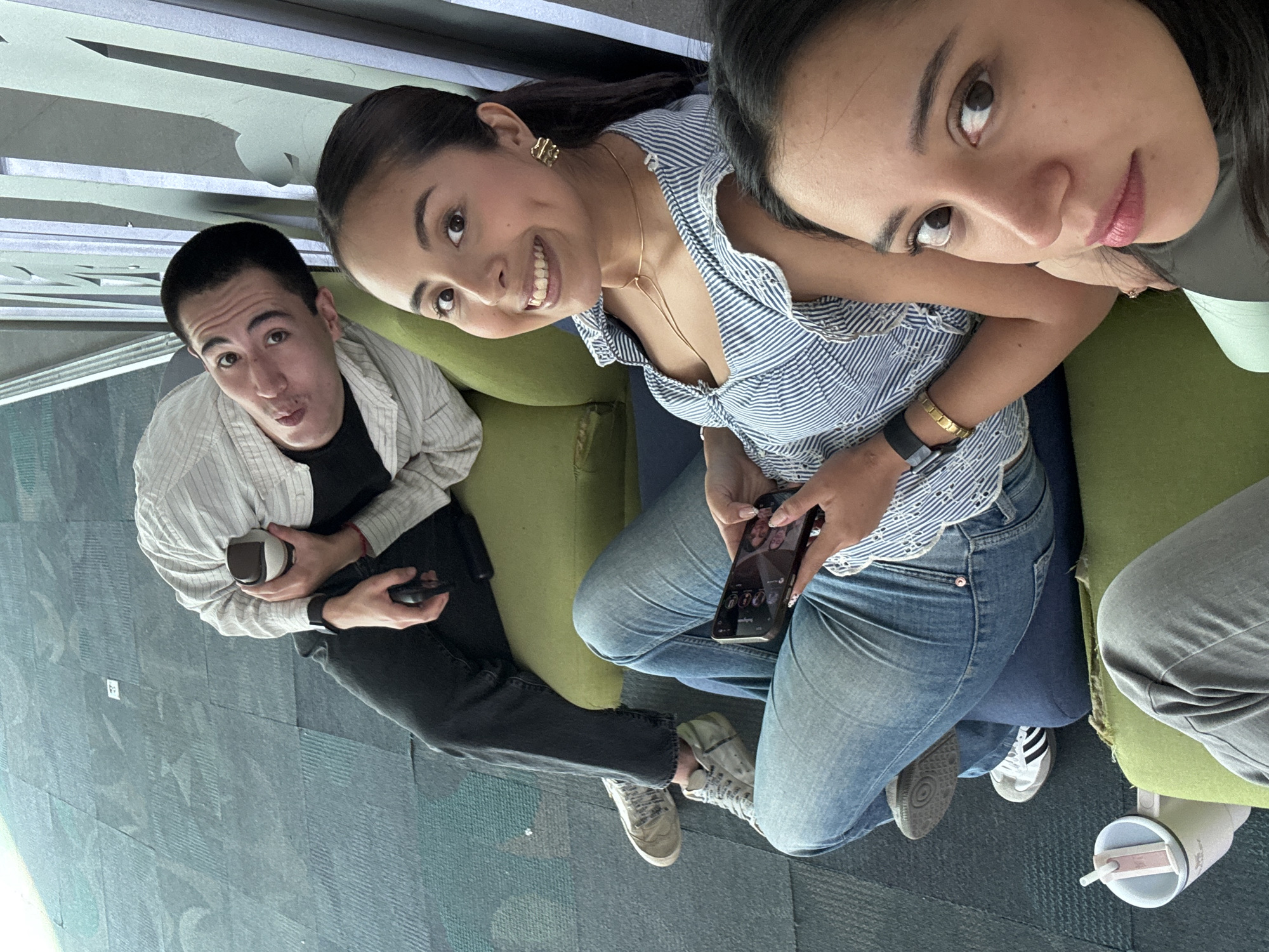 From a high-angle perspective, the photo depicts three people sitting on a green couch. The man on the left, wearing a black t-shirt and striped white jacket, makes a comical face while holding a cup. The woman in the center, wearing a striped blue and white top and blue jeans, smiles and holds a phone. The woman on the right looks at the camera with a neutral expression. The floor is covered in blue carpet tiles. A white cup is positioned near the right. The exact location in Puebla, Mexico, cannot be determined from this image.