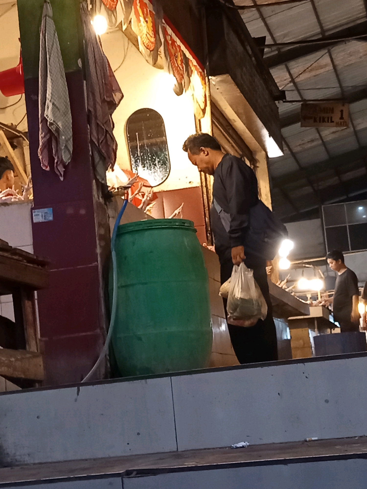 The image depicts an indoor market scene, possibly in Karawang, Indonesia, with a man in the foreground carrying a plastic bag of groceries. He is wearing a dark long-sleeved shirt and dark pants, with a blue bag slung over his shoulder. To his left is a large green barrel and part of a maroon-colored structure. Behind him, a younger man in a dark t-shirt stands near a display of what appears to be seafood on ice. Above the stalls, a corrugated metal roof is visible, with overhead lighting illuminating the area. A sign with Indonesian text is also visible hanging from the ceiling. The overall impression is that of a busy, functional marketplace.