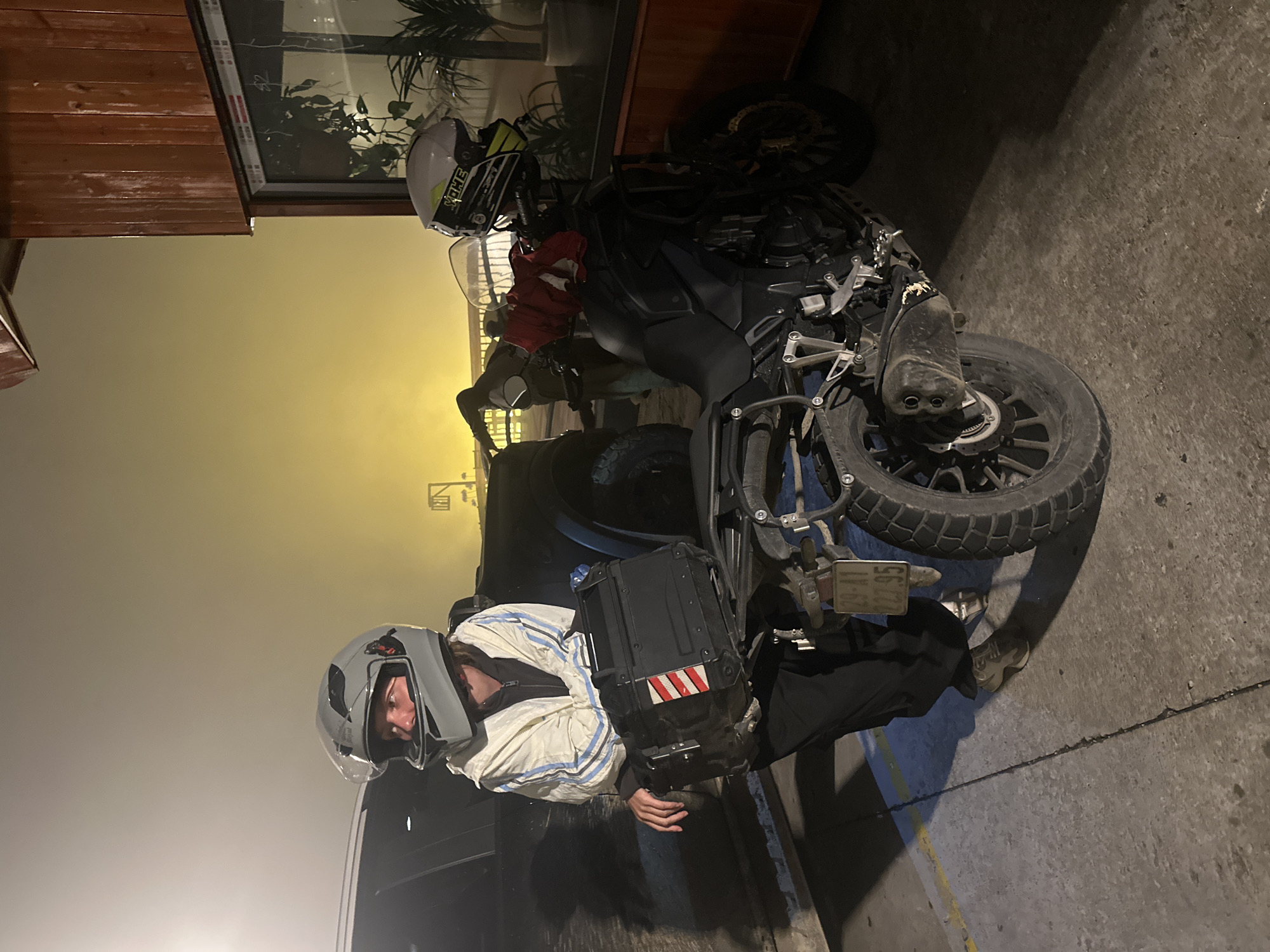 The image shows a person in a motorcycle helmet standing next to a motorcycle. The motorcycle is equipped with a top box. The location appears to be outdoors, possibly a street or parking area, as indicated by the pavement and nearby building. The photograph was taken at night in Binh Lu, Vietnam. The overall lighting is dim.