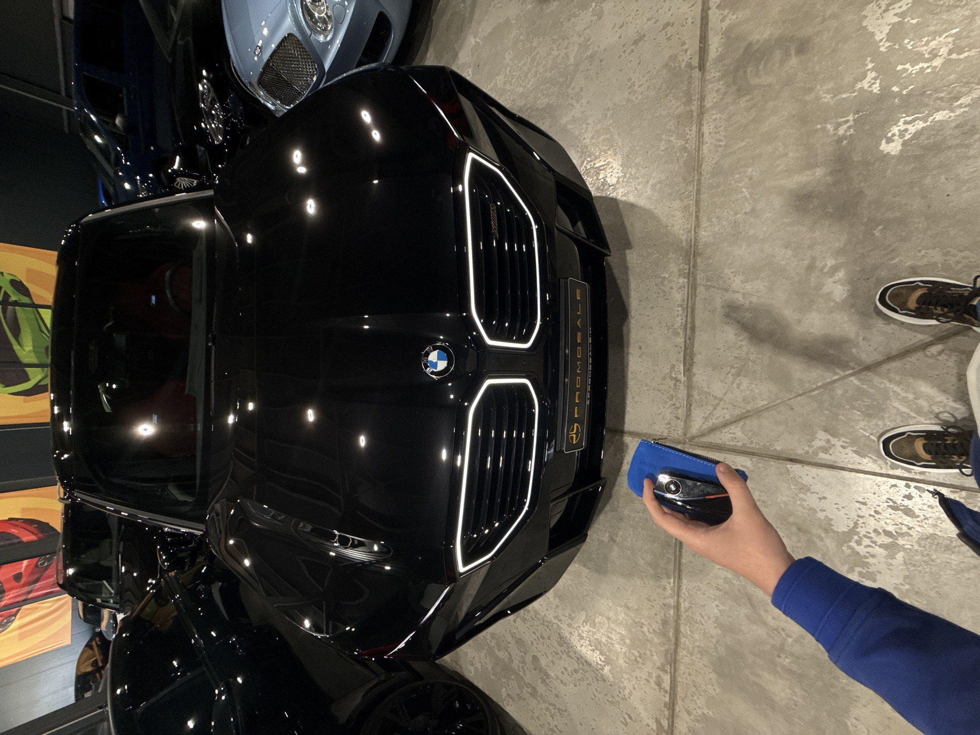 A high-angle shot shows the front of a black BMW vehicle parked indoors on a gray concrete floor. A person wearing a blue long-sleeved shirt holds a blue pouch containing a car key near the car's front grill. The person's shoes, dark brown and white sneakers, are visible. Other luxury vehicles, including a Bentley, are partially visible in the background. The car has a distinctive illuminated kidney grill design with vertical bars outlined in white light. The "PROMOSALE" logo is visible on a plate to the right of the grill. The image appears to have been taken in a luxury car dealership in Plovdiv, Bulgaria.