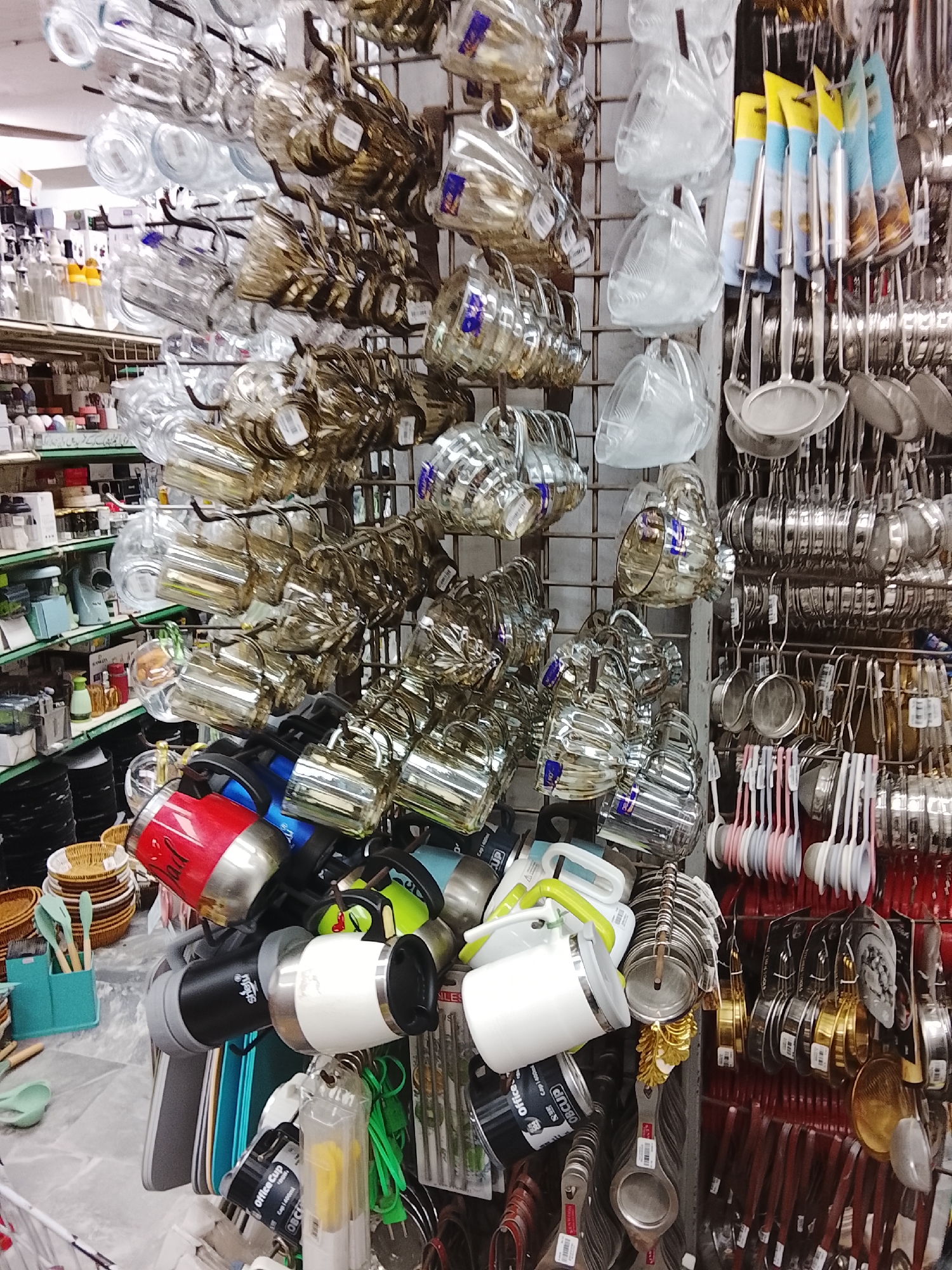 The image shows a cluttered display of kitchenware in a retail setting, likely a shop in Gulgasht Colony, Pakistan. Various types of mugs and cups made of glass and metal hang from a metal grid. Below the mugs, there are metal cooking utensils, including ladles, skimmers, and measuring spoons. The shelves in the background hold containers and other kitchen-related items. The scene shows no people.