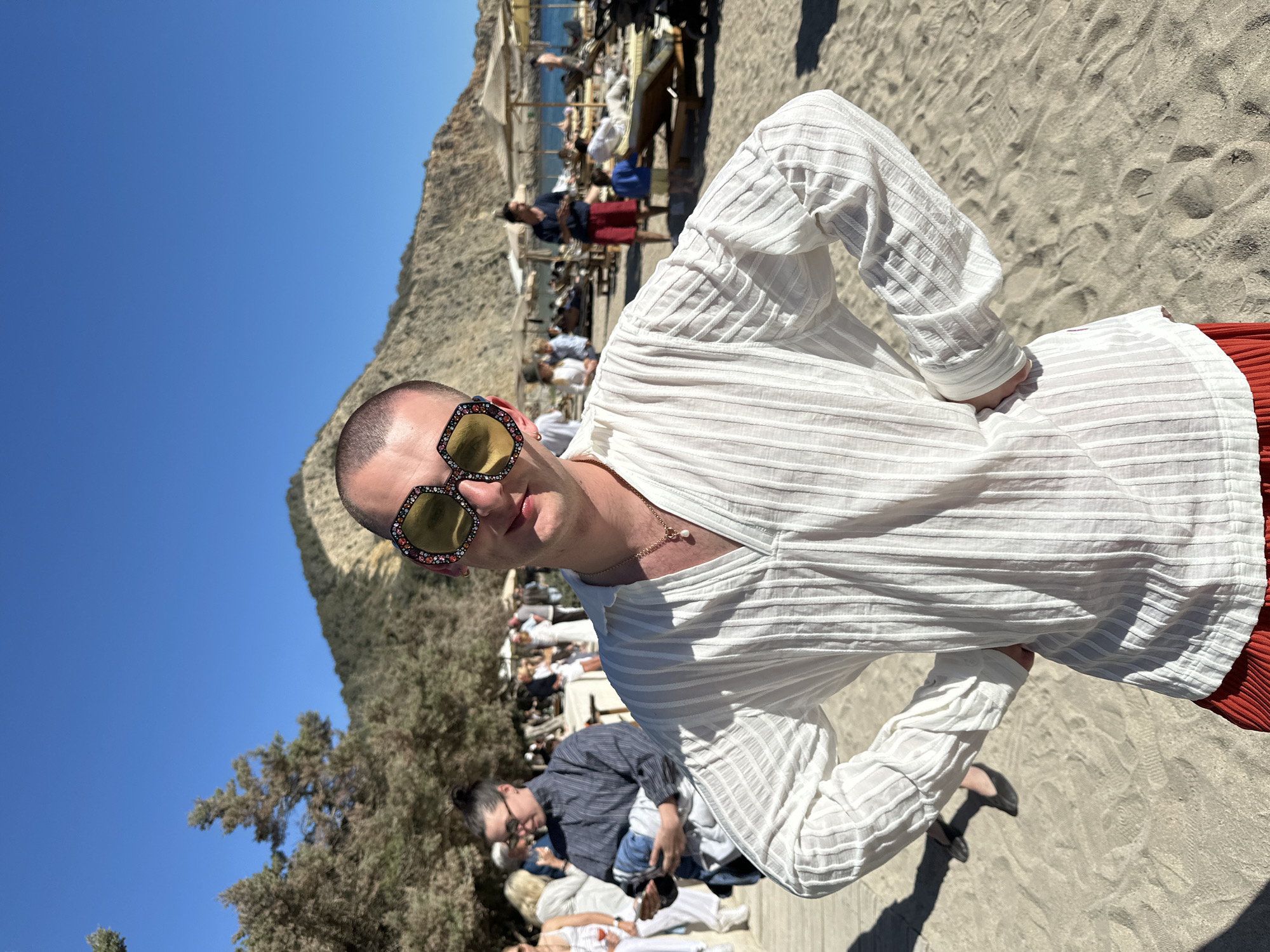 A man with sunglasses and a white striped shirt stands on a sandy beach in Sant Josep de sa Talaia, Spain. He is facing the viewer and has his hands on his waist. Other people are lounging on beach chairs in the background. There are trees, a mountain, and a clear blue sky behind him.