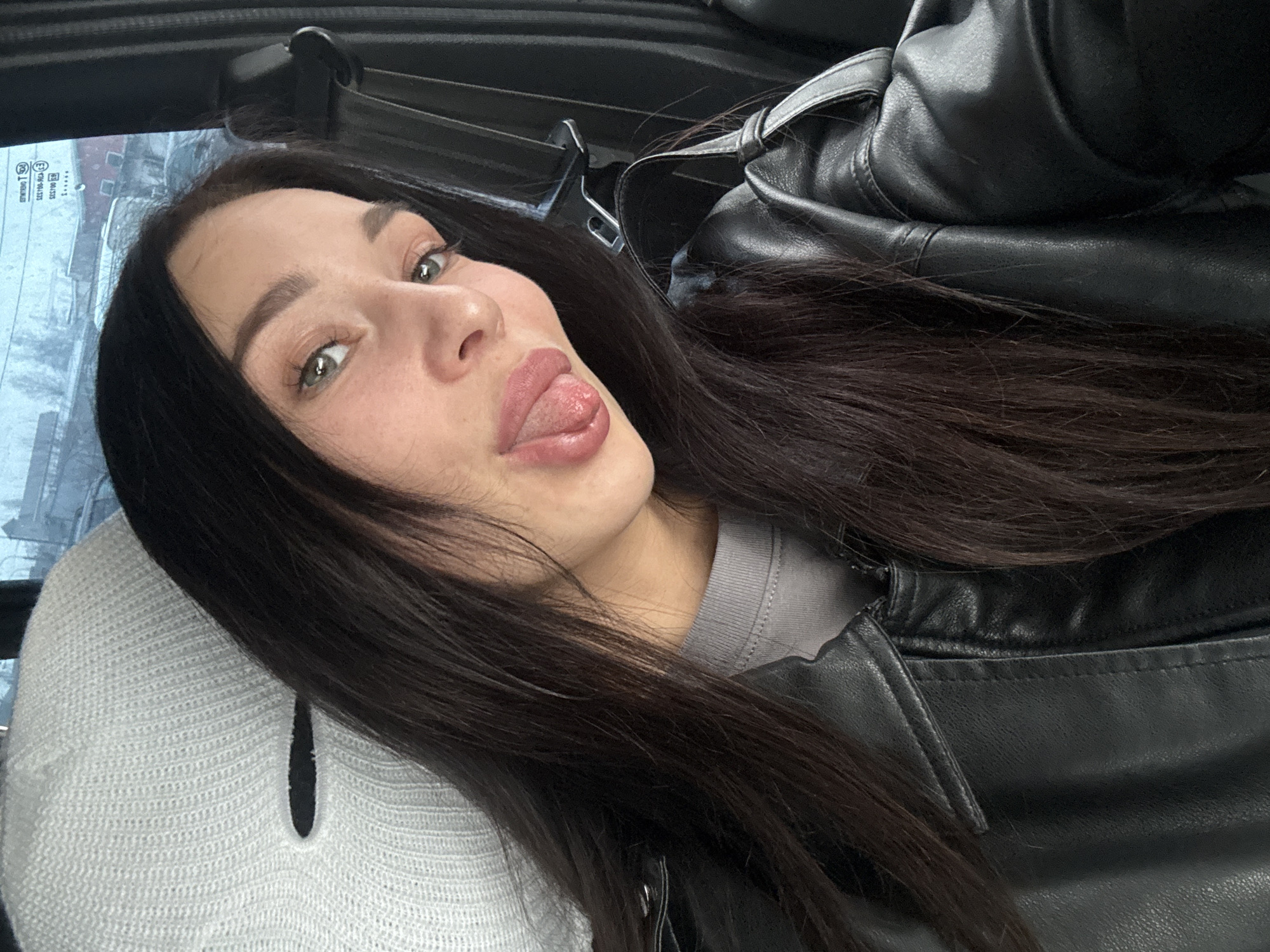 A young woman poses for a selfie, sticking her tongue out playfully. She has dark, straight hair and wears a black leather jacket over a gray top and a white knitted sweater or cardigan. The photo appears to have been taken inside a vehicle, as indicated by the window and the car interior in the background. The window reveals a view of Chaikovsky, Russia, with buildings and possibly snowy or overcast conditions visible outside. A portion of the car's interior can be seen at the top of the image.