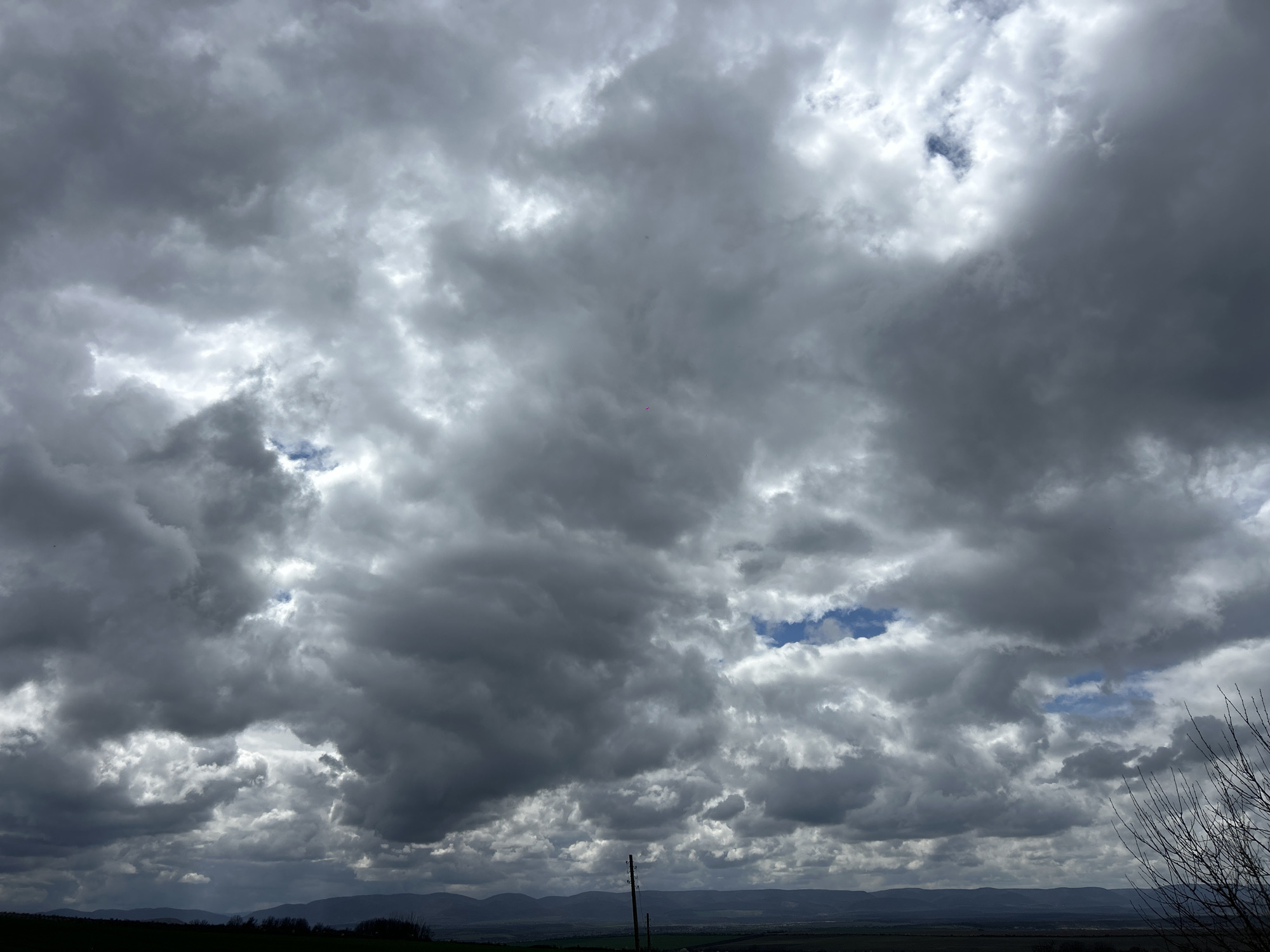The image depicts a cloudy sky over a landscape. The sky is dominated by heavy, gray clouds with occasional patches of blue visible through openings. The landscape below features a rolling terrain of green fields and distant hills. There is a utility pole visible in the lower portion of the image, and some bare branches on the right side. The scene suggests a rural setting. There are no people visible in the image. The location is not explicitly stated in the image to be Srednia, Bulgaria, but is assumed based on external data.