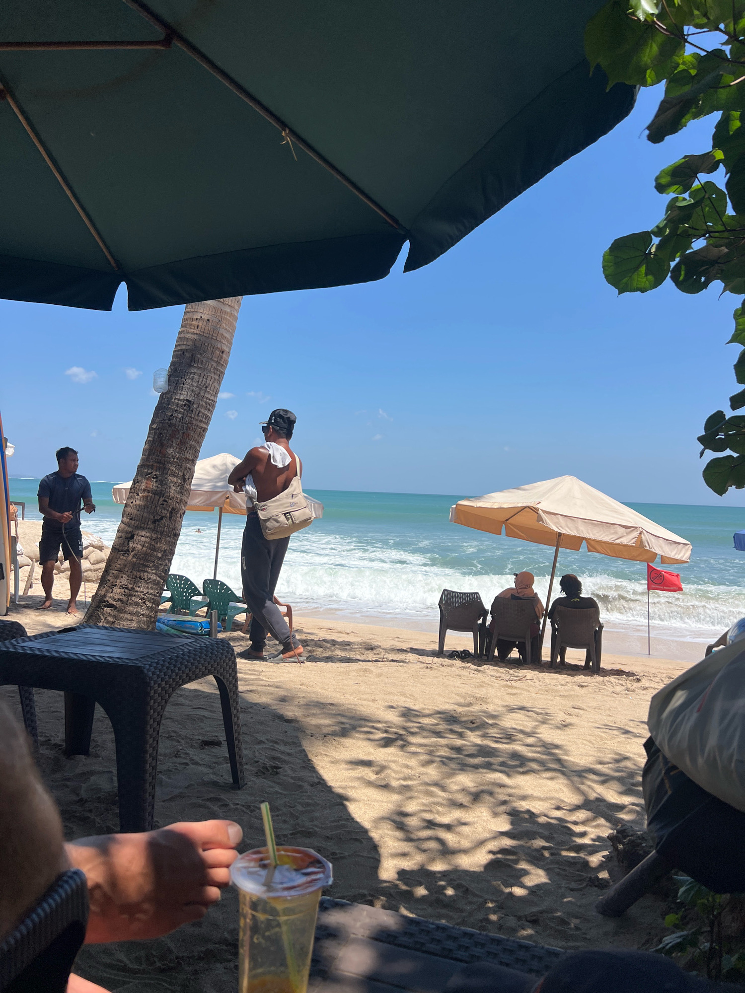 The image depicts a beach scene in Kuta, Indonesia, under a bright, sunny sky. In the foreground, a person's foot rests near a plastic cup with a straw and an orange-colored beverage. A woven-style table is also visible in the foreground.
Further back on the sandy beach, two men are standing near a palm tree. One man, wearing a dark t-shirt and shorts, holds a dark rope. The other man, shirtless and wearing a baseball cap, has a light-colored bag slung over his shoulder and a towel draped around his neck. A beach umbrella provides shade behind them.
In the middle ground, three people are seated in chairs under a beige umbrella, facing the ocean. Two individuals are closely seated together, and another person is seated to their left. A red flag with a graphic symbol is planted in the sand near the umbrella. The ocean is calm with gentle waves breaking on the shore. In the distance, the horizon is visible with clear blue sky above. The overall atmosphere is relaxed, suggesting a vacation or leisure setting.