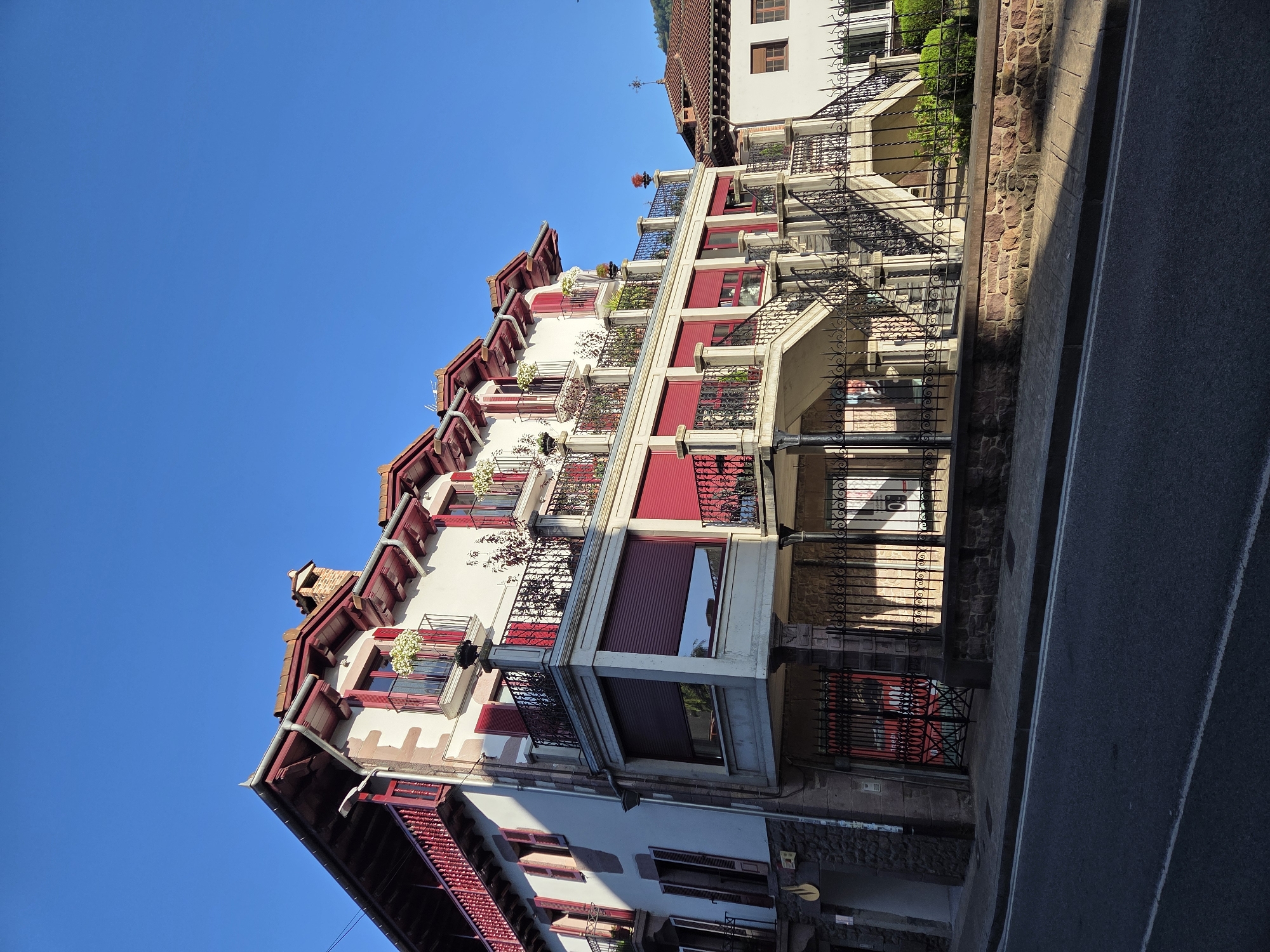 A multi-story building with a white facade and red accents is shown in the city of Baztan, Spain. The building features several balconies with ornate black railings and red shutters. A grand staircase with black metal banisters leads up to the second story. The street in front of the building is paved and bordered by a low stone wall. The sky is clear and blue.