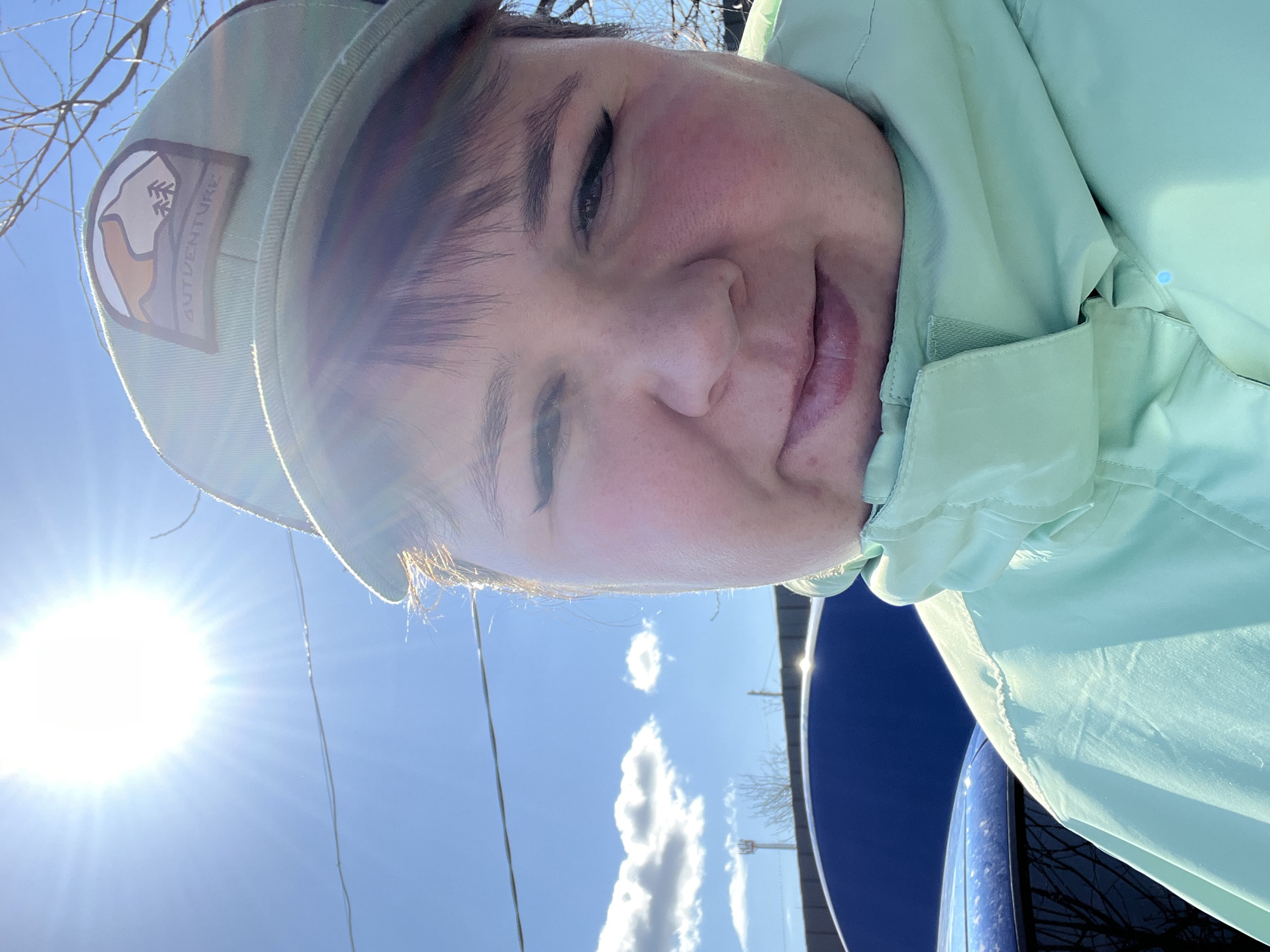 A selfie, likely taken in Chaikovsky, Russia. A person is wearing a light green jacket and matching cap with an "Outventure" patch. The background features a clear blue sky with some clouds and bare branches, suggesting early spring. The sun is shining brightly, causing lens flare. A dark blue object, possibly a vehicle, is visible in the lower right. The person is looking slightly downward with a small smile.