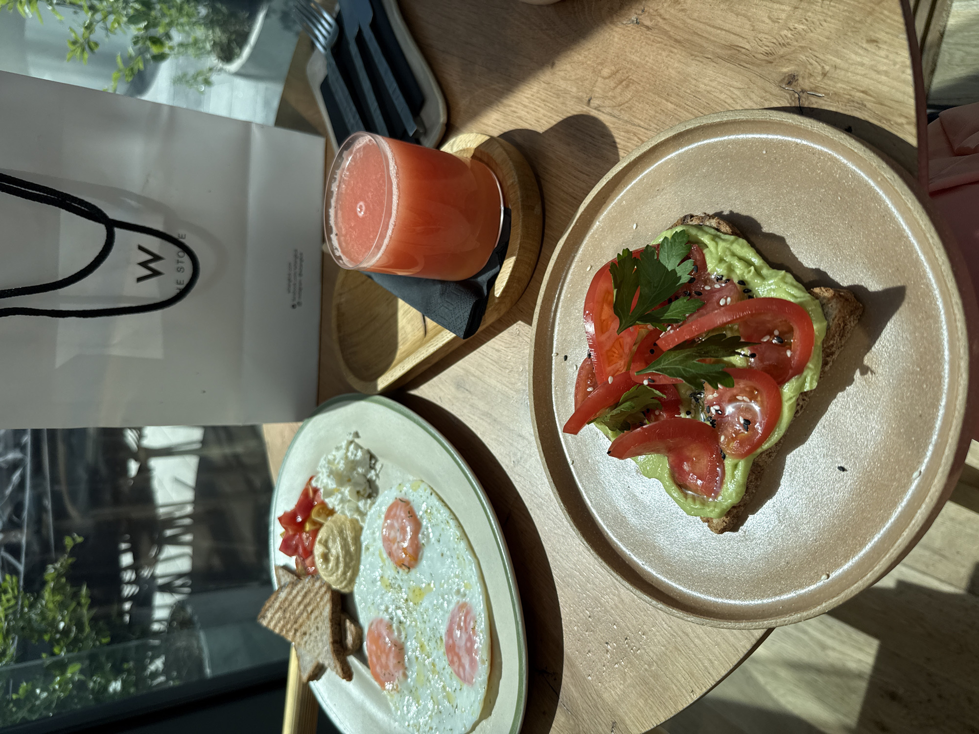 The image captures a dining table scene, likely in a cafe or restaurant setting. In the foreground, two plates of food are visible. One plate holds avocado toast topped with sliced tomatoes and herbs, possibly parsley. The second plate contains a fried egg dish with what appears to be cottage cheese, tomatoes, and toast. A glass of pink juice rests on a wooden coaster, accompanied by a black napkin. A shopping bag with the letter "W" printed on it is partially visible in the background, suggesting a retail purchase. The location appears to be in Khimki, Russia, based on the logo on the bag. Natural light streams through a nearby window, creating shadows and highlighting the textures of the food and the wooden table. Utensils are placed on a wooden serving tray. The overall setting suggests a casual and modern dining experience.