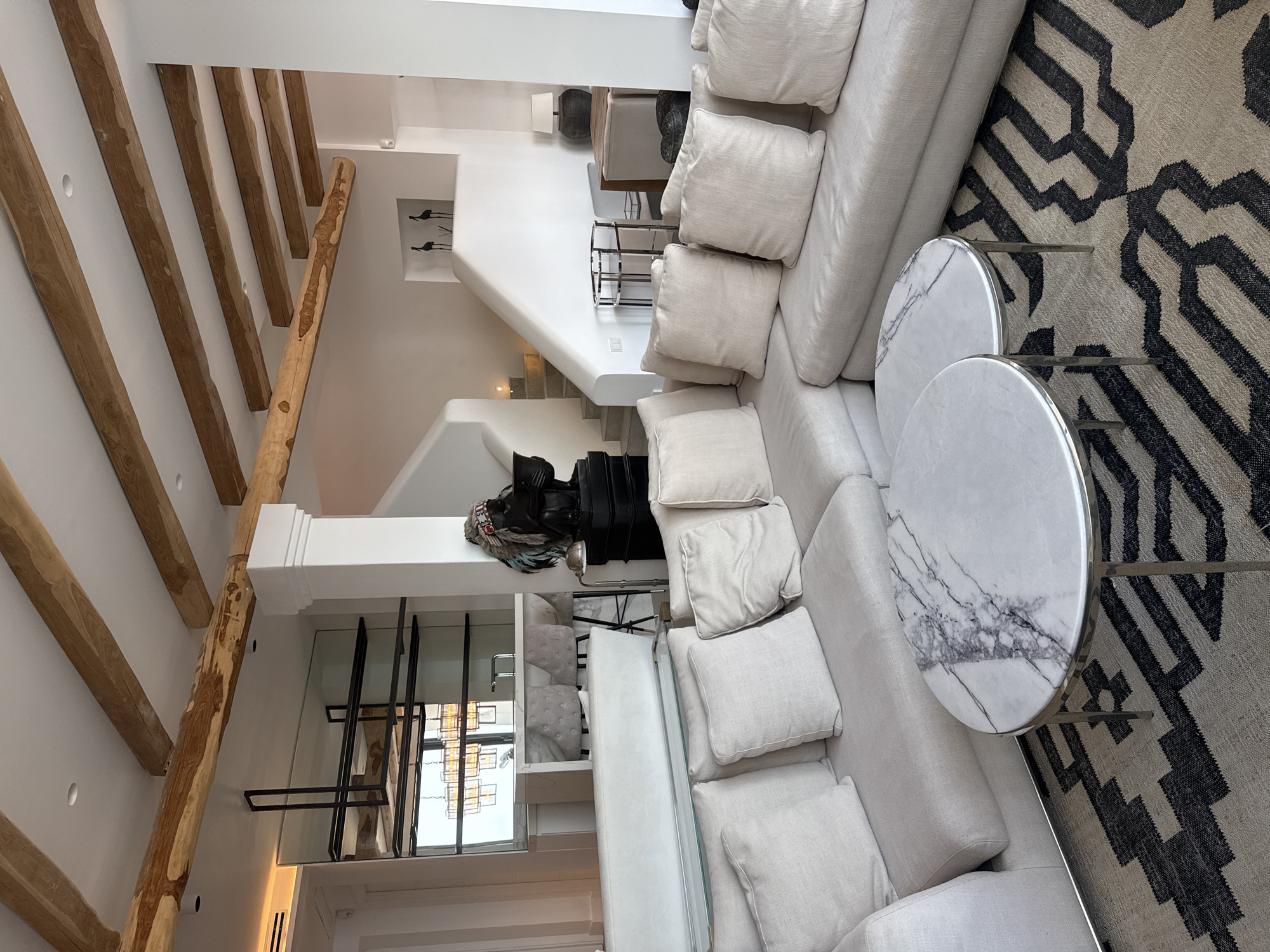 This image depicts a modern, open-plan living space, likely a lounge area in a high-end property, potentially a villa or hotel suite, in Santa Eulària des Riu, Spain. The room features a large, light-colored sectional sofa with numerous plush cushions arranged around a rug with a geometric black and beige pattern. Two round coffee tables with white marble tops and metallic bases are placed in front of the sofa.
The ceiling is a prominent feature, with exposed, rough-hewn wooden beams adding a rustic, natural element that contrasts with the clean, white walls. A minimalist bar area with mirrored backsplash, sleek shelving, and bar stools is visible in the background, suggesting a connection to a kitchen or entertainment space. Further back, a white, curved staircase leads to an upper level.
Decorative elements include a dark statue of a figure wearing a headdress, positioned near the staircase, and a few smaller decorative items like ceramic birds on a shelf. A floor lamp with a metallic shade stands beside the statue. The overall aesthetic is a blend of modern luxury and natural textures, creating a sophisticated and comfortable atmosphere. The lighting appears to be a mix of ambient and task lighting, highlighting the architectural features and furnishings.