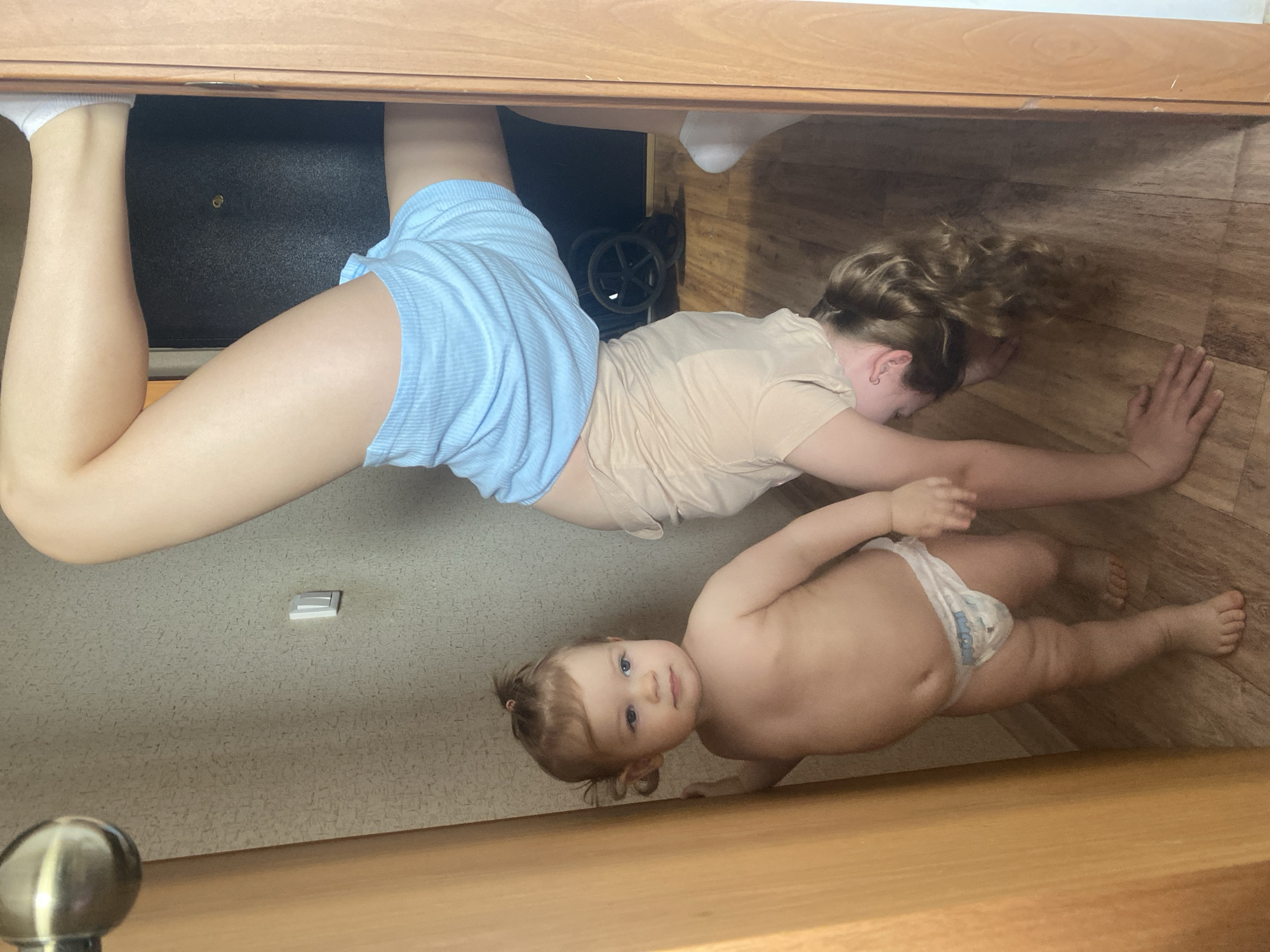 A low-angle, full-length shot depicts two children in what appears to be a makeshift play area. A taller child wearing light blue shorts and a tan top is oriented horizontally at the top of the frame, with her head reaching toward the right corner and her feet extending towards the upper-left corner. Beneath her, a baby in a diaper looks towards the camera. The scene is lit by overhead lighting that casts shadows on the walls and floor. The "play area" appears to be formed by the underside of a wooden structure, possibly a piece of furniture. The background includes what appears to be a wooden floor.