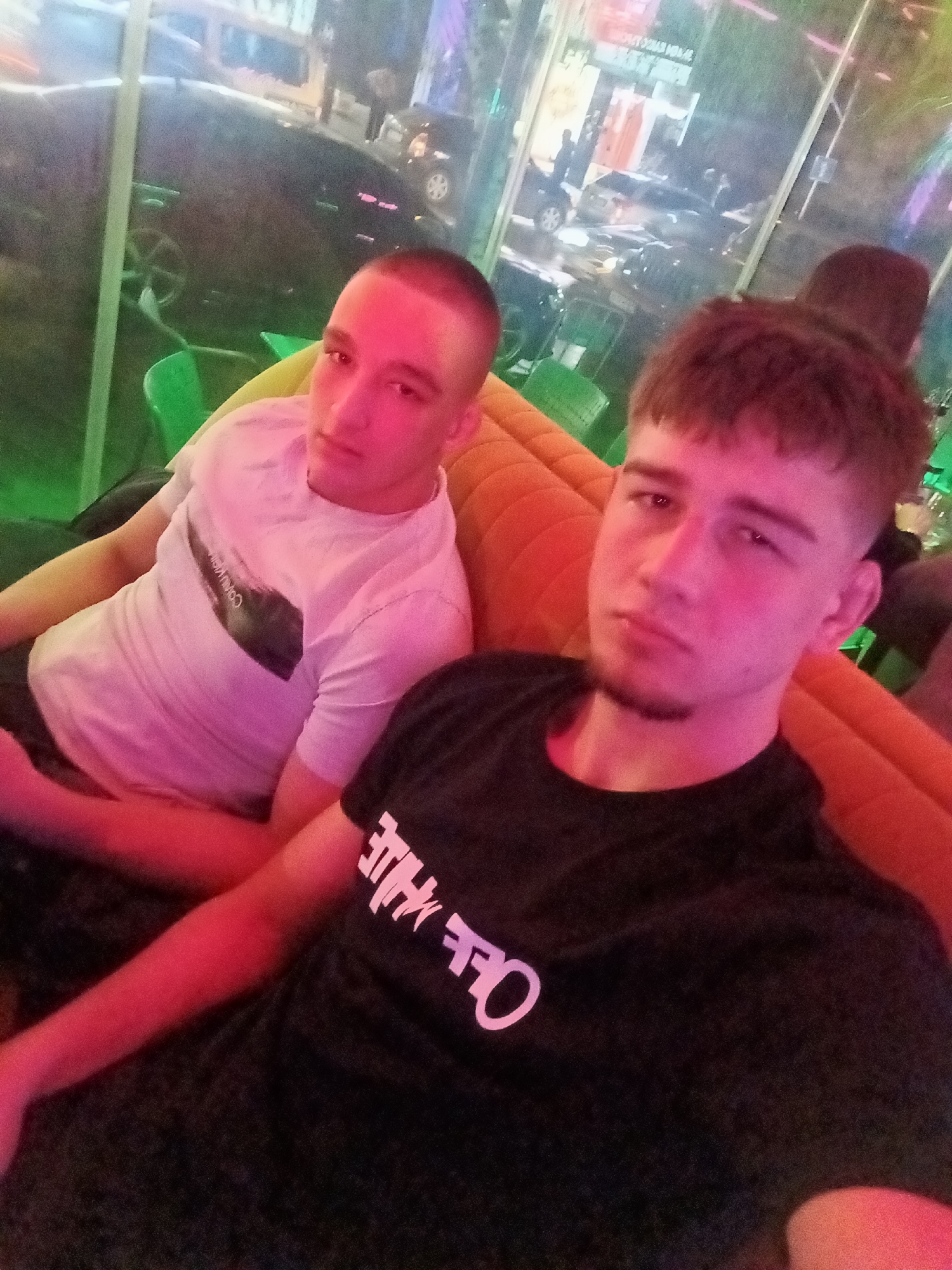 Two young men are seated on an orange couch inside a room. The man on the left wears a white t-shirt. The man on the right wears a black t-shirt with white lettering. The background shows a window with reflections of vehicles and lights. The location appears to be an indoor space in Sofia, Bulgaria.