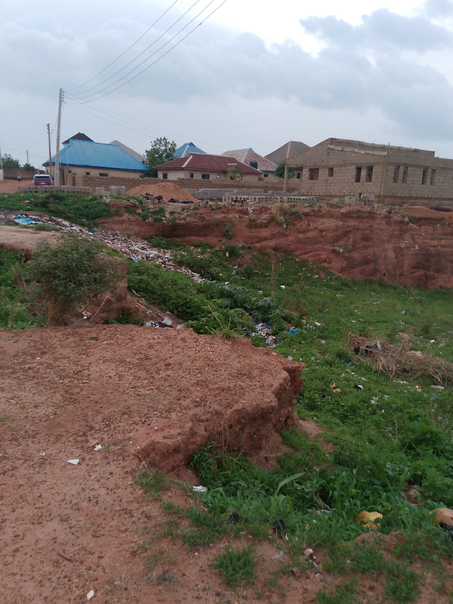 The scene depicts a drainage ditch in Barakin Angundi, Nigeria, filled with trash and debris. The ditch is overgrown with green vegetation and has eroded red dirt banks. Several buildings are visible in the background, including structures with blue roofs and an unfinished block building. Overhead power lines stretch across the sky. A red car is visible in front of one of the buildings. The sky is overcast.