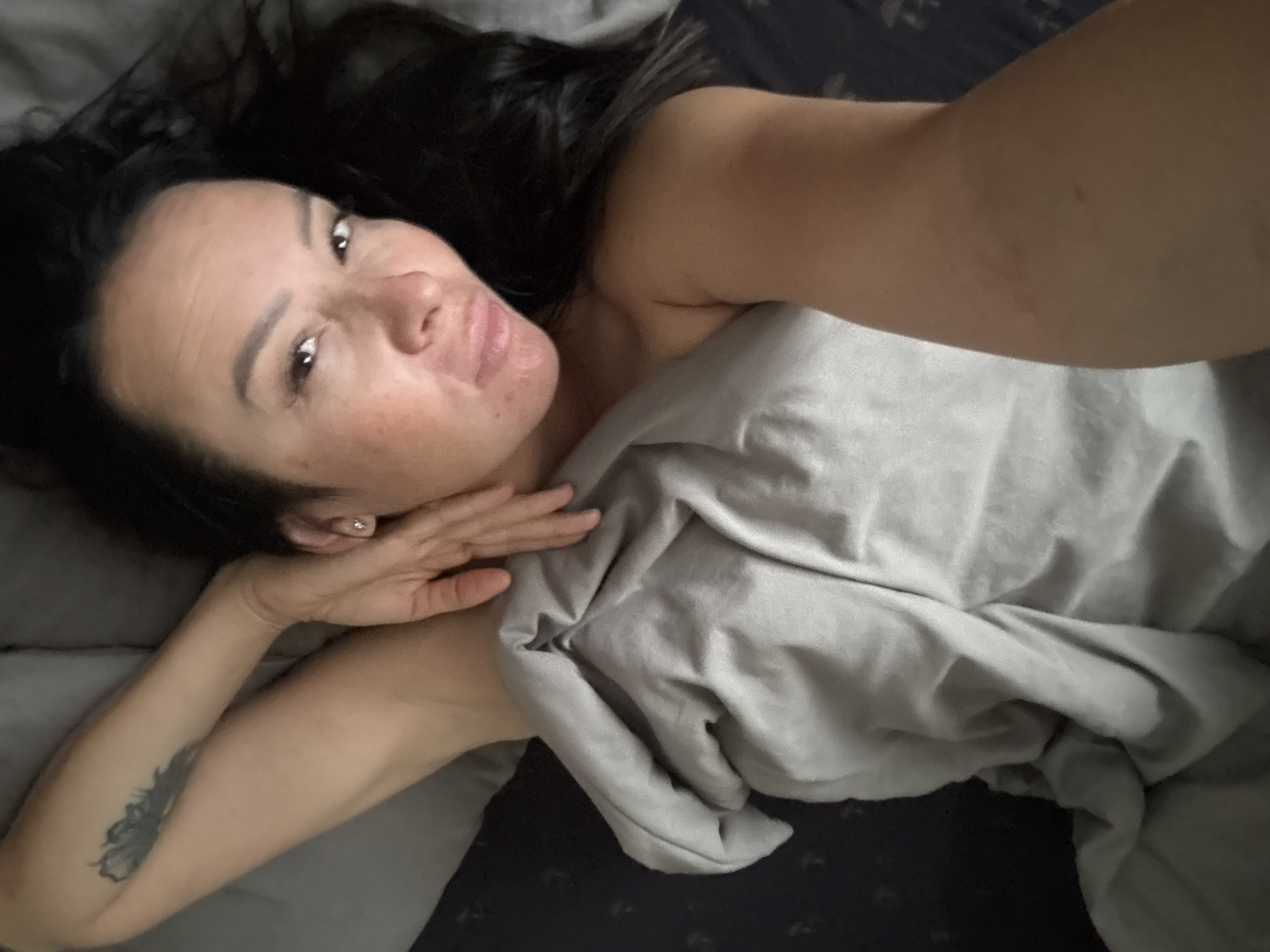 A woman is lying in bed, looking at the camera. She has dark hair and is wearing a diamond stud earring. Her left arm is raised, and she has a tattoo of a feather on her upper arm. She is covered by a grey blanket, and the room appears to be dimly lit.
